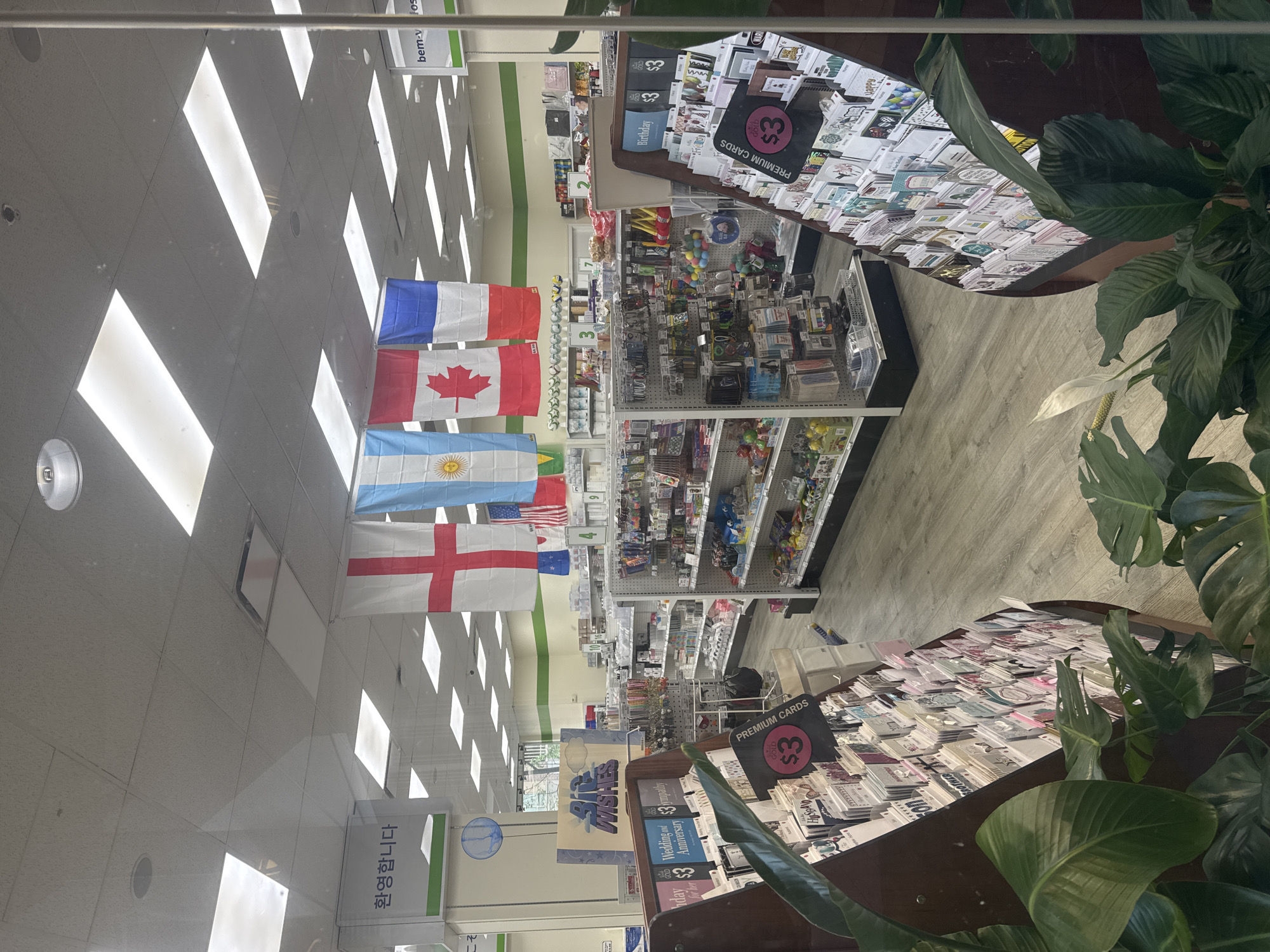 The image shows the interior of a retail store. In the foreground, a display of greeting cards is visible, with a plant in front of it. The store shelves are stocked with various merchandise. Several national flags, including those of England, Argentina, Canada, and France, are hanging from the ceiling. A sign that reads "환영합니다" (Welcome in Korean) is also visible. In the background, more shelves and aisles of products are present. The store appears to be well-lit with fluorescent ceiling lights. No people are clearly visible in the image. The location is not specified in the image itself.