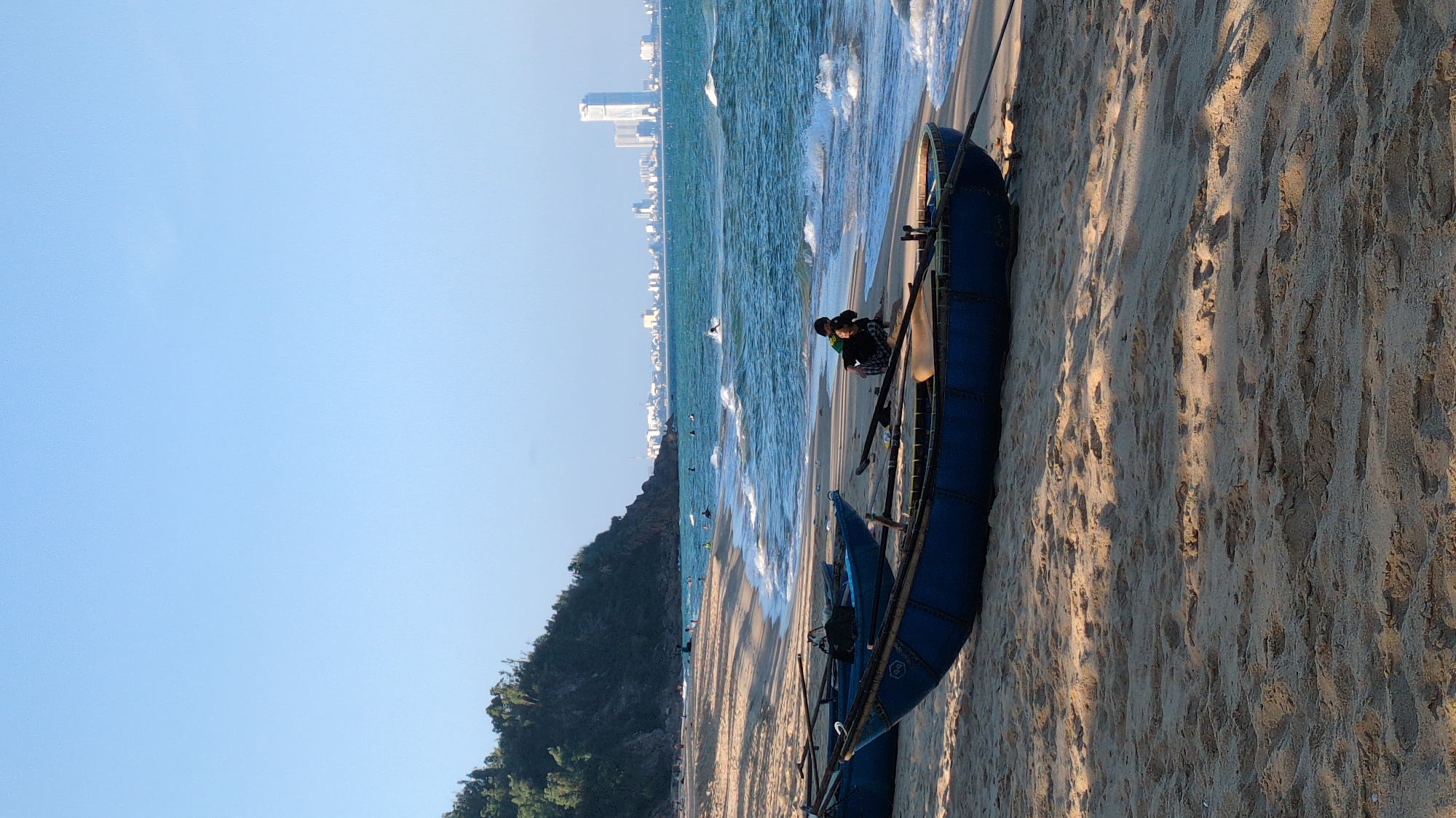 The image captures a coastal scene in South Quy Nhon Ward, Vietnam. Two blue coracle boats are positioned on the sandy beach. Two people sit near the boats. The ocean's edge meets the shore, with visible waves. In the distance, a city skyline is visible. A hill covered in vegetation is situated to the left of the shoreline. The sky is clear and blue. Shadows are cast across the sand, indicating sunlight.