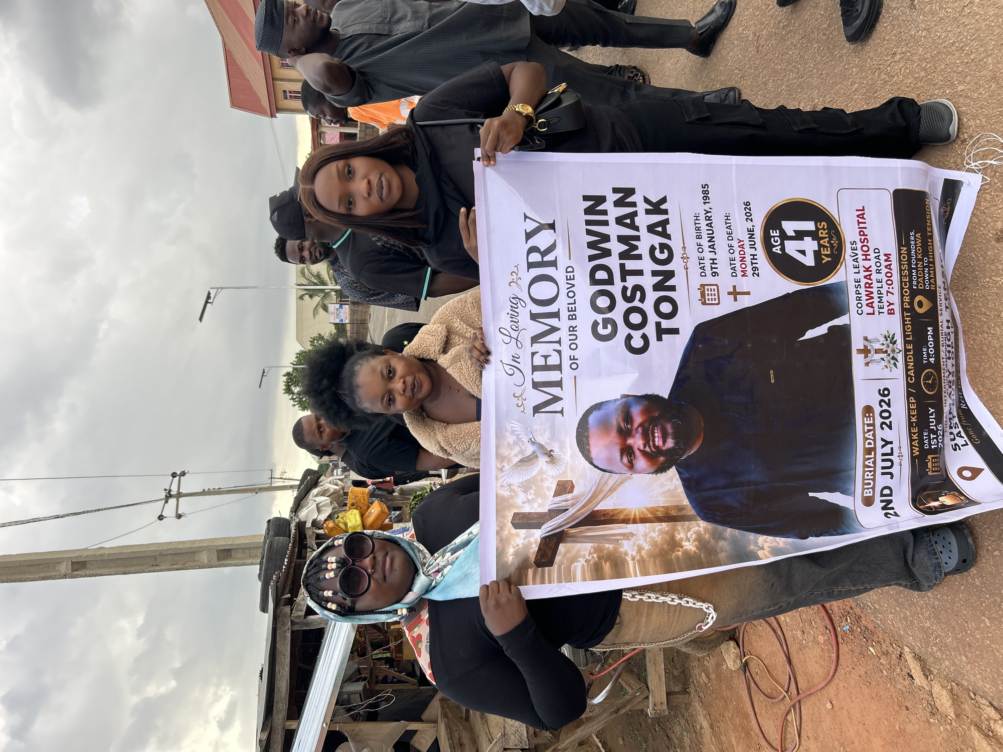 The image shows a group of people in Jos, Nigeria, holding a large funeral banner. The banner features a portrait of a man named Godwin Costman Tongak, who passed away on June 29, 2026, at the age of 41. The banner details his date of birth as January 9, 1985, and provides information about his burial date, wake-keep, and candle light procession. The corpse was to leave Lawrak Hospital Temple Road by 7:00 AM, and the wake-keep was scheduled for July 1, 2026, at 4:00 PM. The burial is set for July 2, 2026.
The people in the image appear somber, reflecting the solemnity of the occasion. The setting seems to be outdoors, possibly on a street or in a community area, with a cloudy sky suggesting it might be a humid or overcast day. Some structures and utility poles are visible in the background, hinting at an urban or semi-urban environment. The overall mood conveyed is one of mourning and remembrance for the deceased.