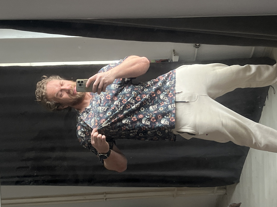 A single male subject is depicted indoors, visible through a mirror selfie. The specific location within Saint Petersburg, Russia, cannot be determined from the visual information provided.
The subject is an adult male with light brown, wavy hair and a beard. He is wearing a dark t-shirt with a repeating pattern of light-colored skulls and pink and blue floral designs. His lower garment consists of light-colored (possibly off-white or light grey) sweatpants with a dark drawstring and a small dark logo on the left hip. A dark-banded wristwatch is visible on his left wrist.
He is holding a light-colored smartphone in his right hand, angled to capture his reflection in the mirror. His left hand is raised, with the index finger extended and pointing towards the fabric of his t-shirt on his chest. The subject's eyes appear partially closed or squinting, and his mouth is slightly open.
The background visible in the reflection features a dark, possibly black or grey, curtain behind the subject on the left side. Partially visible to the far left are white vertical radiator pipes. On the right side of the reflection, a segment of a light-colored wall is present, along with a vertical dark panel and a small white shelf-like object. The floor appears to be a light, uniform surface.