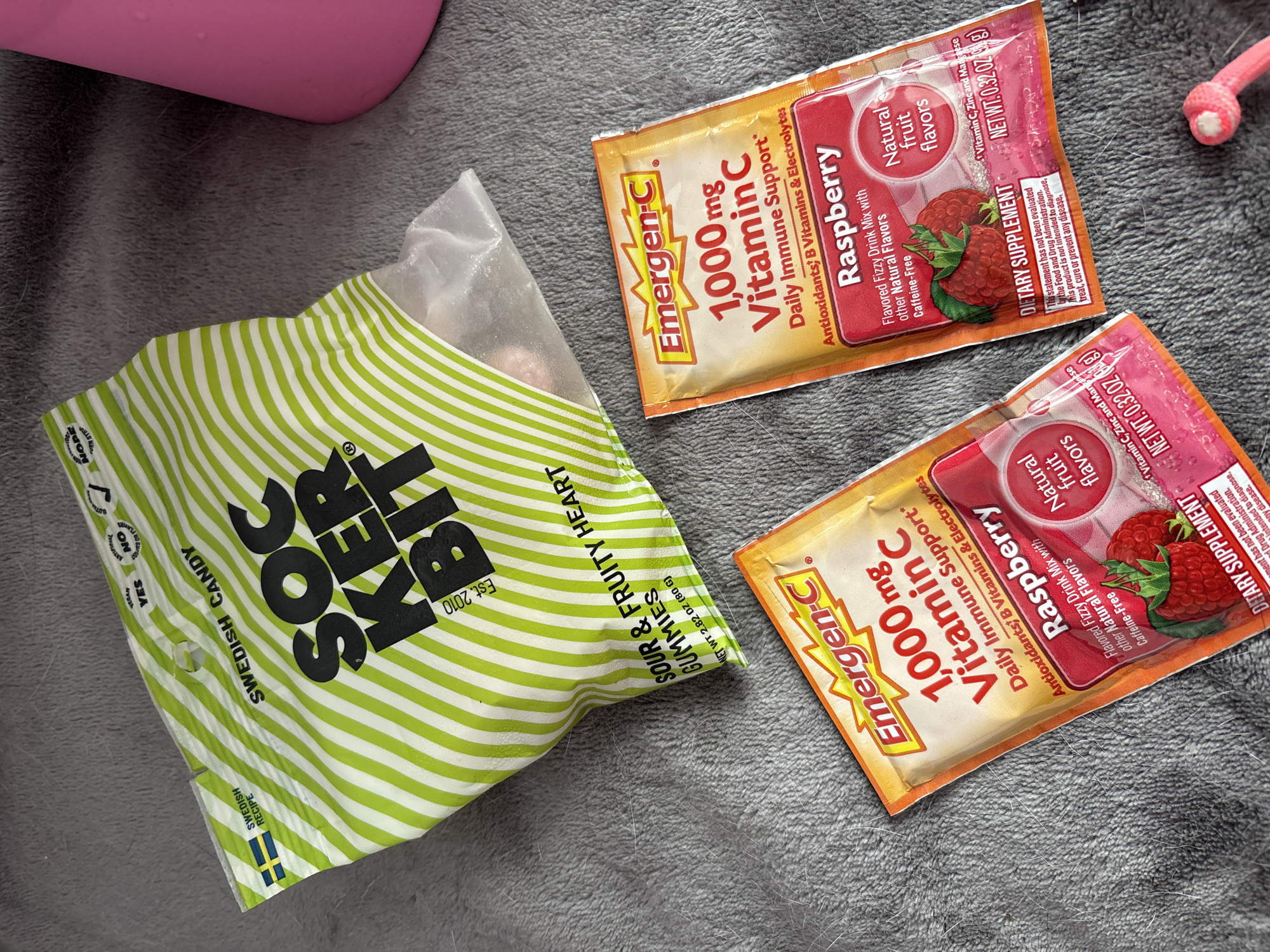 The image shows a gray surface, possibly a blanket or rug, on which several items are placed. There's a green and white striped bag of "Sockerbit" Swedish candy. Next to it are two packets of "Emergen-C" vitamin C drink mix in raspberry flavor. The packets are orange with red accents and display information about the product. The bag of candy is labeled "Sour & Fruity Heart Gummies". A pink object and a pink object with white details, are also visible. The scene does not provide enough information to specify a location in Seal Beach, United States of America.