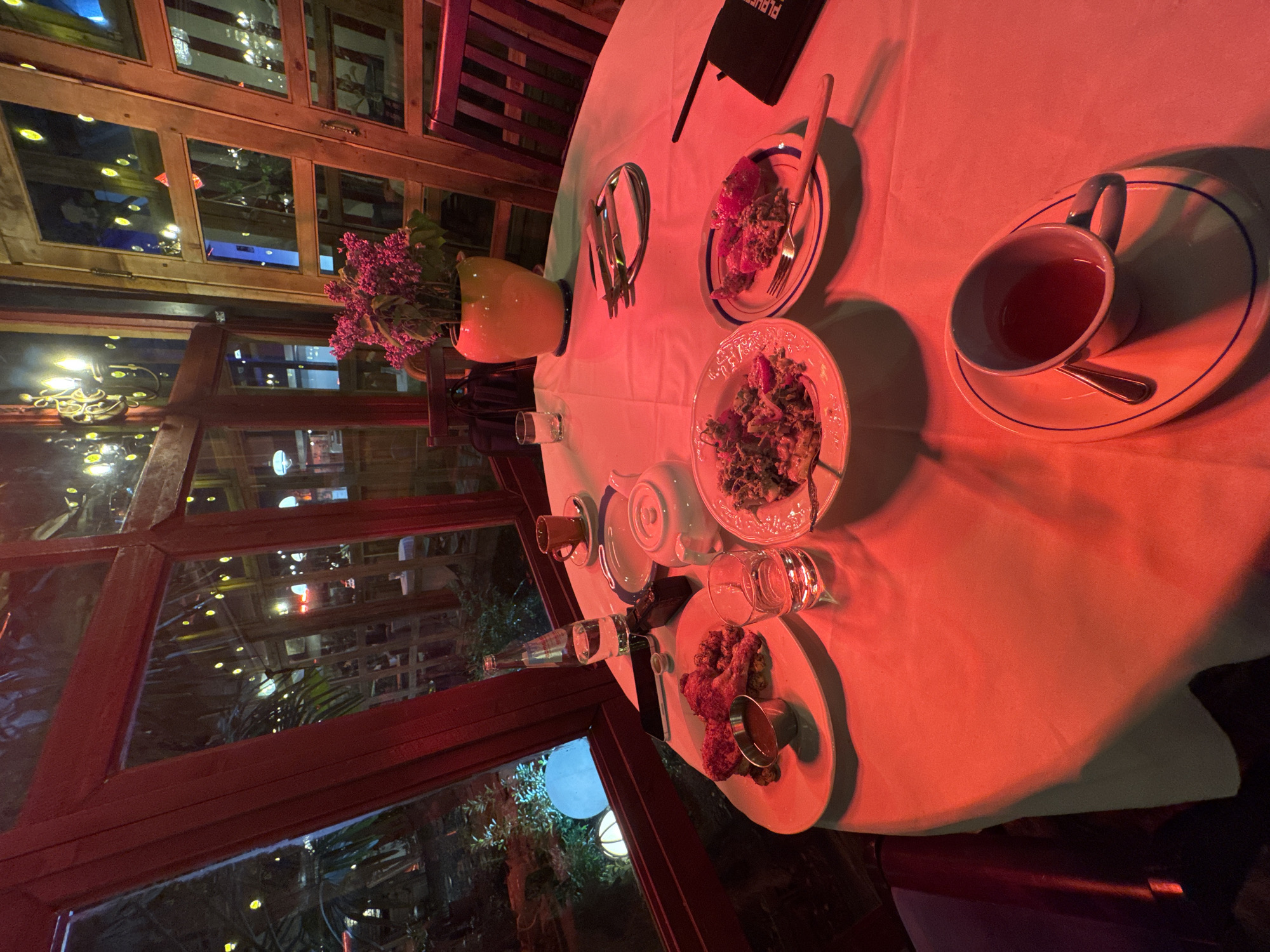 The image captures an intimate dining scene at night inside what appears to be a restaurant or cafe in Tbilisi, Georgia. The entire space is bathed in a strong, warm red and orange ambient light, creating a cozy and somewhat dramatic atmosphere.
In the foreground, a round table covered with a white tablecloth is set with several dishes and drinks. On the table, there are multiple plates holding various food items, including what looks like salads or appetizers and some fried components with a small dipping sauce cup. Teacups and saucers are present, one containing a dark liquid (likely tea) with a spoon, and another smaller cup and teapot. Glasses of water, one accompanied by a water bottle, are also visible. A small yellow vase holds a bouquet of purple flowers, possibly lilacs. Cutlery, specifically forks, is placed near the plates. A dark menu or stand, partially visible, has white text that begins with "PLU...". A smartphone also lies on the table.
The table is positioned next to large windows with wooden frames, offering a view of an outdoor area. Through the windows, reflections of the indoor lights mix with the view of an outdoor space, which features string lights and at least one round, glowing white light fixture, suggesting a garden or patio. No people are visible in the frame, but the table setting indicates it is prepared for at least two diners. The scene suggests an evening meal or tea session in a warmly lit, inviting establishment.
