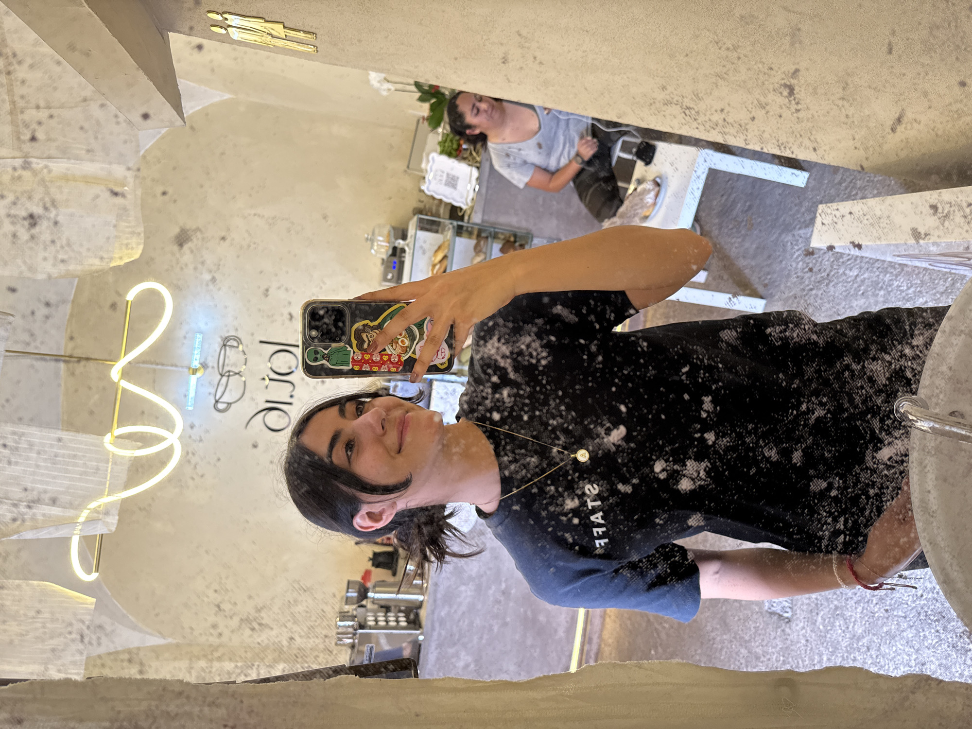 A young woman is captured in a mirror selfie, smiling gently as she holds her smartphone. She is wearing a black long-sleeved shirt with the word "STAFF" (reversed) printed on it, and a delicate gold necklace with two small circular charms, one bearing the letter 'A' and the other 'V'. Her phone case is adorned with several colorful stickers, including an alien, a cat, and what appear to be Russian dolls. A red string bracelet is visible on her right wrist.
The most striking detail is the pervasive white, powdery substance coating the woman's shirt, arms, and hair, as well as parts of the mirror's surface, suggesting an activity involving flour or powdered sugar, or a decorative effect.
The setting appears to be a cafe or bakery. In the background reflection, another woman is visible, sitting or working at a counter, wearing a light-colored t-shirt. A coffee machine, a display case with pastries or bread, and a QR code are also reflected. A white neon sign on the wall, spelling out "lorie" (reversed) with a coffee cup logo, further confirms the cafe ambiance. The walls have a mottled, textured appearance, and decorative gold-colored human figures are mounted on an upper section of the wall. The overall scene is well-lit, suggesting an indoor environment during operational hours. The image is taken in Puebla, Mexico.