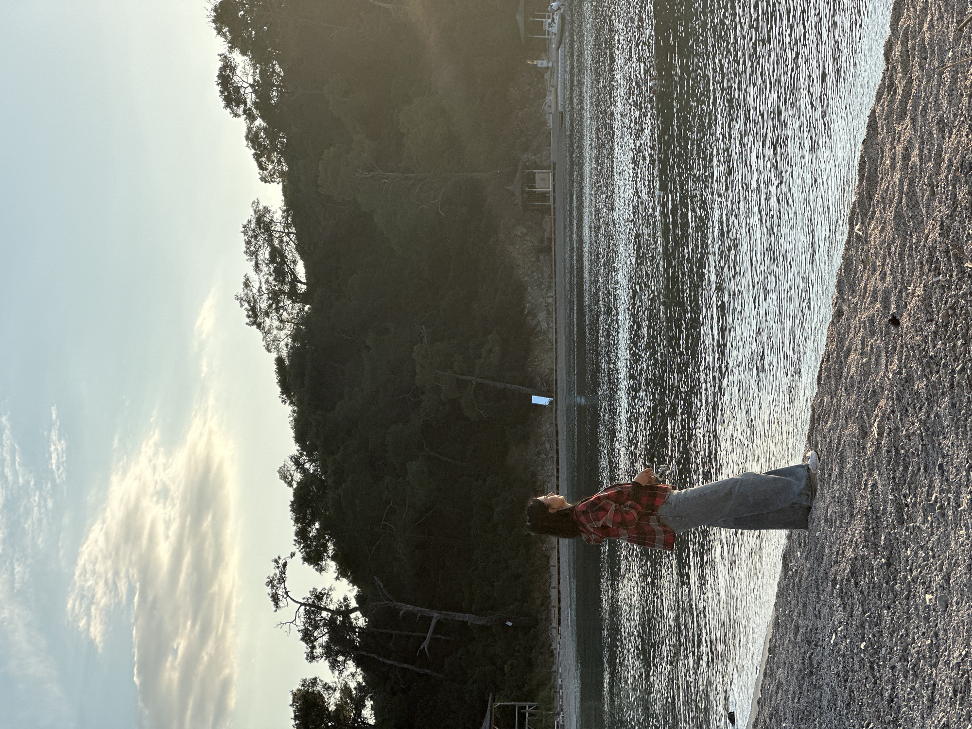 A woman stands on a pebble beach alongside calm, rippled water, likely the sea, looking up at a forested hillside. She is wearing a red and black plaid shirt and blue jeans with white sneakers. The sky is pale with some clouds. The location appears to be a bay or cove. The city of Fethiye, Turkey is not discernible in this image.