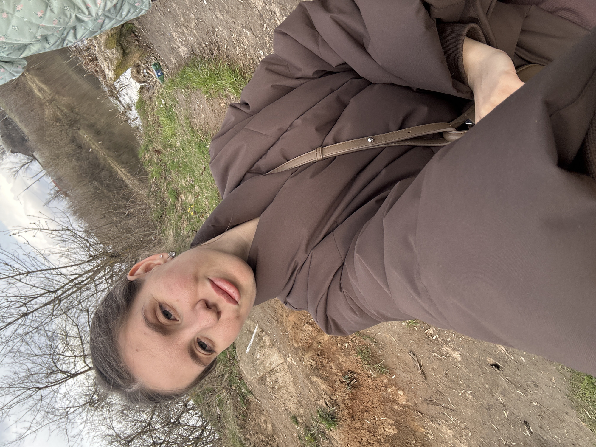 The image shows a person in a brown puffer jacket and cross-body bag standing in an outdoor area near a body of water. The ground appears to be dirt and grass with sparse vegetation. Bare trees are visible in the background. Based on the location provided, this scene is likely in or near Klin, Russia.