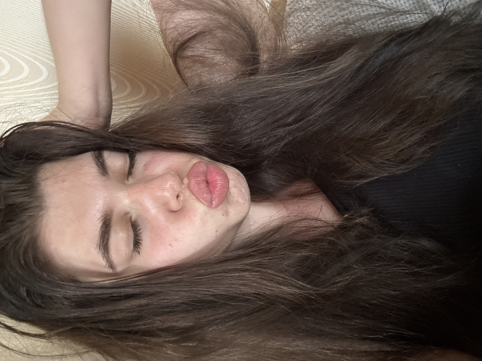 The image depicts a woman with long, brown hair lying down. Her eyes are closed, and her lips are pursed in a kissing gesture. She is wearing a black top. The background appears to be a textured surface, possibly bedding or furniture. No evidence indicates that this image was taken in the city of Мещёры, Russia.