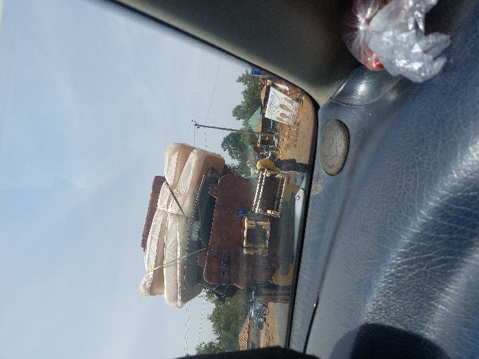 The image depicts an outdoor scene in Hoss, Nigeria, viewed from the interior of a vehicle. The primary subject is a dark-colored truck, positioned ahead on an unpaved, reddish-brown road. The truck is overloaded with household goods, specifically a tall stack of light-colored mattresses, individually wrapped in clear plastic and secured with dark straps or ropes. Beneath the mattresses are several darker, rectangular furniture components, likely disassembled sofa frames. A single individual, potentially a female, wearing a yellow headscarf and darker clothing, stands near the truck's rear on the right side. The background features a clear blue sky, scattered green foliage, and several rudimentary buildings or market stalls constructed from earth-toned materials and corrugated metal roofs. A utility pole with power lines is visible on the right. A large, partially obscured white banner with colorful graphics stands in the background to the far right. The immediate foreground shows a dark, textured vehicle dashboard with a circular speaker grille and a clear plastic bag containing red items on the far right.