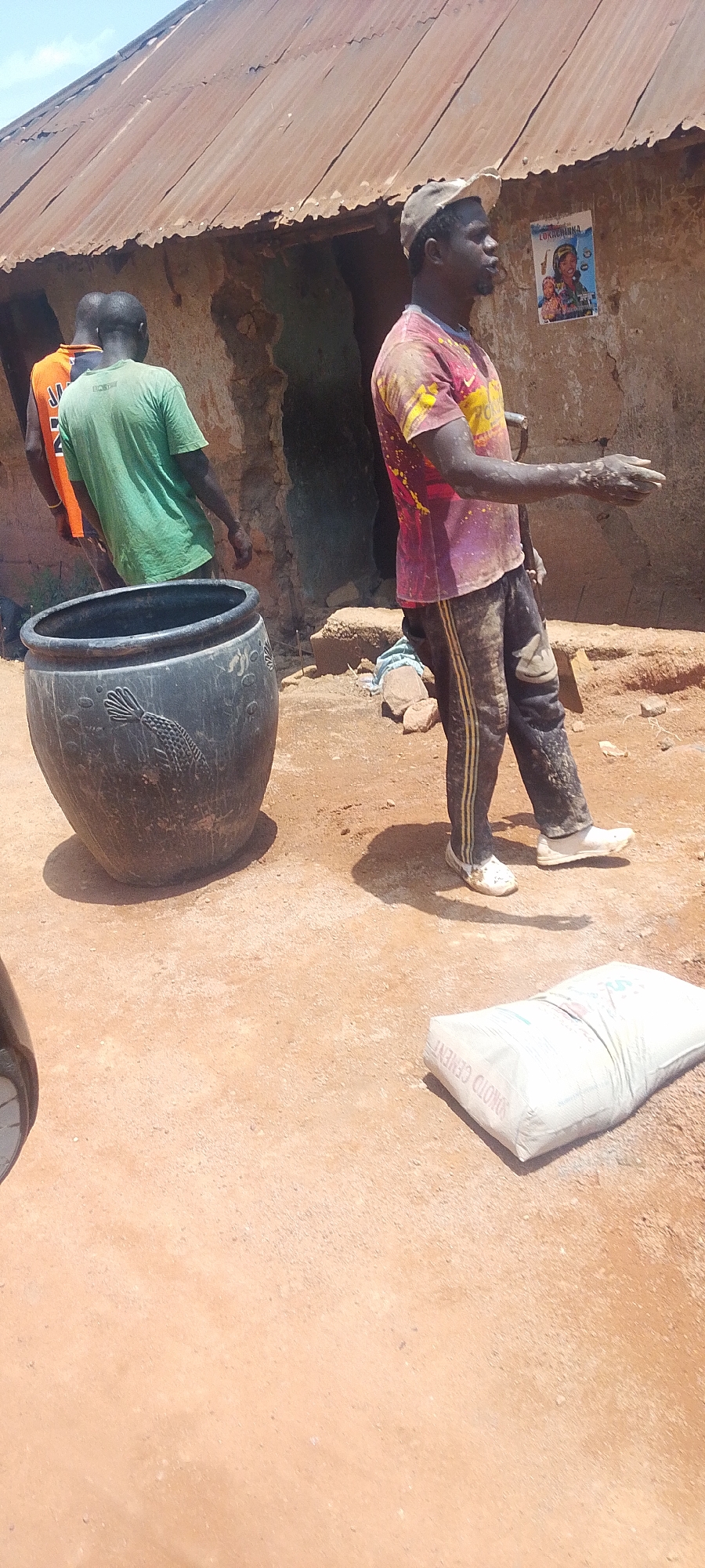 The image depicts a scene in a dusty, outdoor environment, likely a construction or work site, in the city of Jos, Nigeria. Three men are visible. One man in the foreground, wearing a dirty pink and yellow tie-dye t-shirt and dark pants with yellow stripes, stands with his left arm outstretched. He wears a light-colored baseball cap. To his left, a large, dark, round object, possibly a pot or container, rests on the ground. Behind these two men, another man is visible from the back, wearing a green t-shirt. Further to the left, a third man, partially obscured, wears an orange shirt. The background features a rustic building with a corrugated metal roof and textured walls. A poster with an image of a woman is affixed to the wall. In the lower right of the frame, a white bag labeled "SONOTO CEMENT" lies on the ground. The ground itself is composed of reddish-brown dirt and scattered debris. The overall atmosphere suggests an active work environment under daylight.
