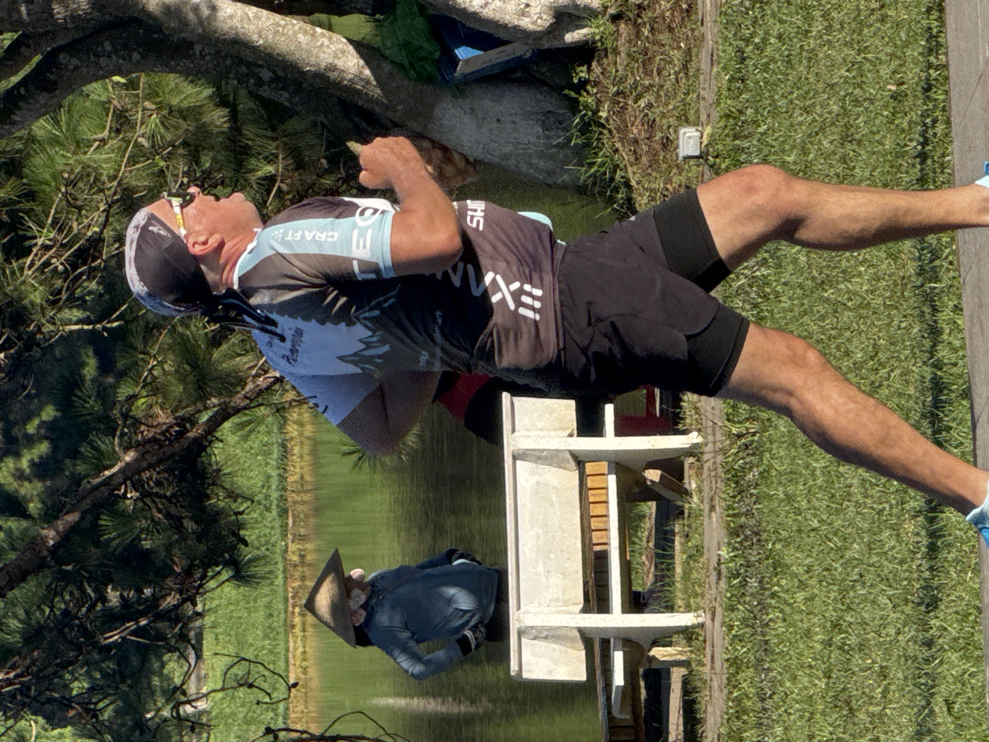 The image depicts a man running outdoors in Da Lat, Vietnam. He is wearing athletic attire, including a bandana, sunglasses, a cycling jersey with the text "CRAFT" visible, and black shorts. In the background, a person wearing a conical hat and a light blue jacket stands by a lake. There is a white bench and some trees present in the scene, suggesting a park or lakeside setting. The environment appears to be sunny with green grass and water.