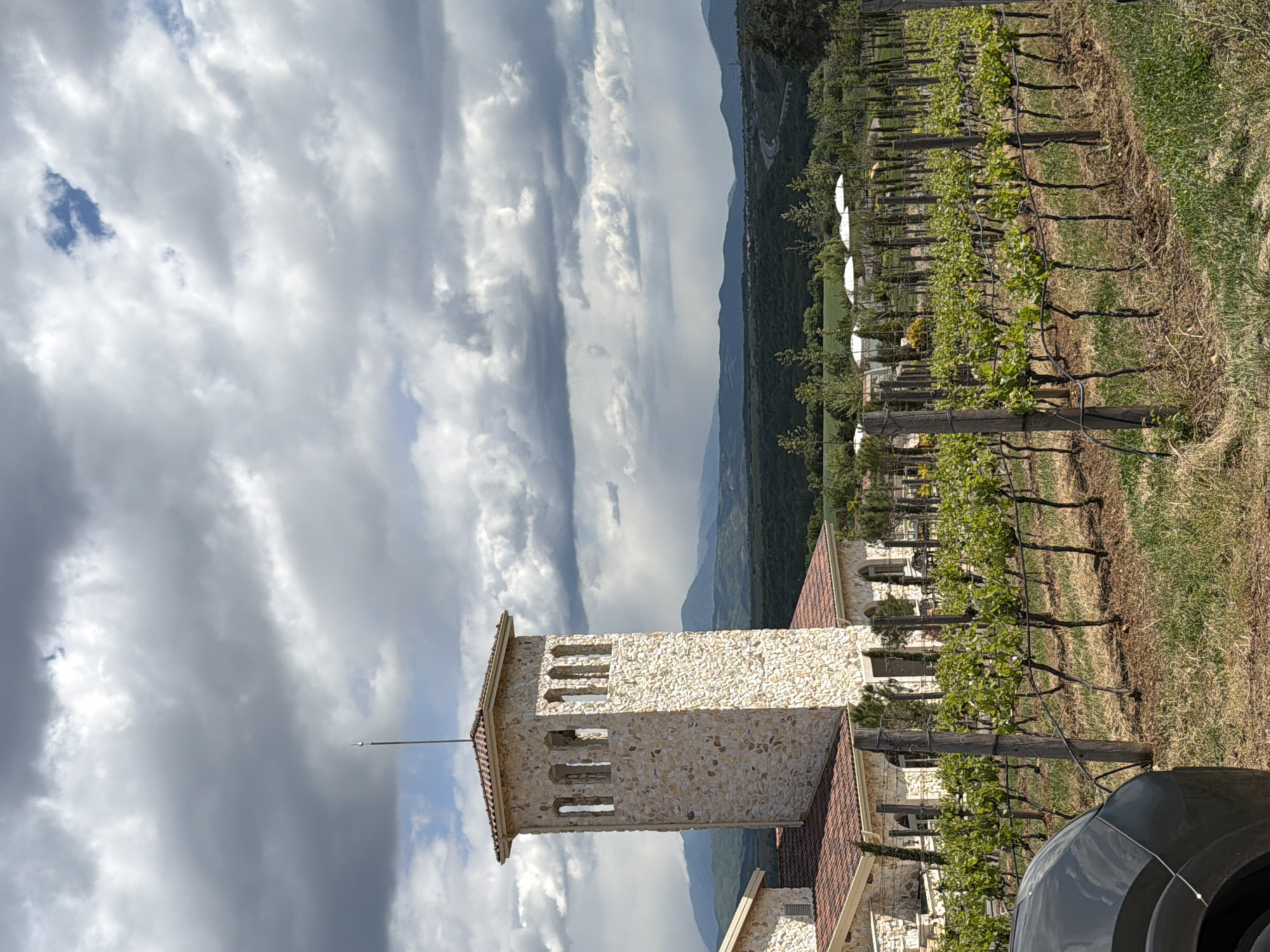 A stone tower with windows is the focal point, positioned near a vineyard with rows of grapevines. The sky is filled with predominantly gray clouds. Distant mountains are visible in the background. Part of a black vehicle is in the lower right corner. This scene is located in Kocherinovo, Bulgaria.