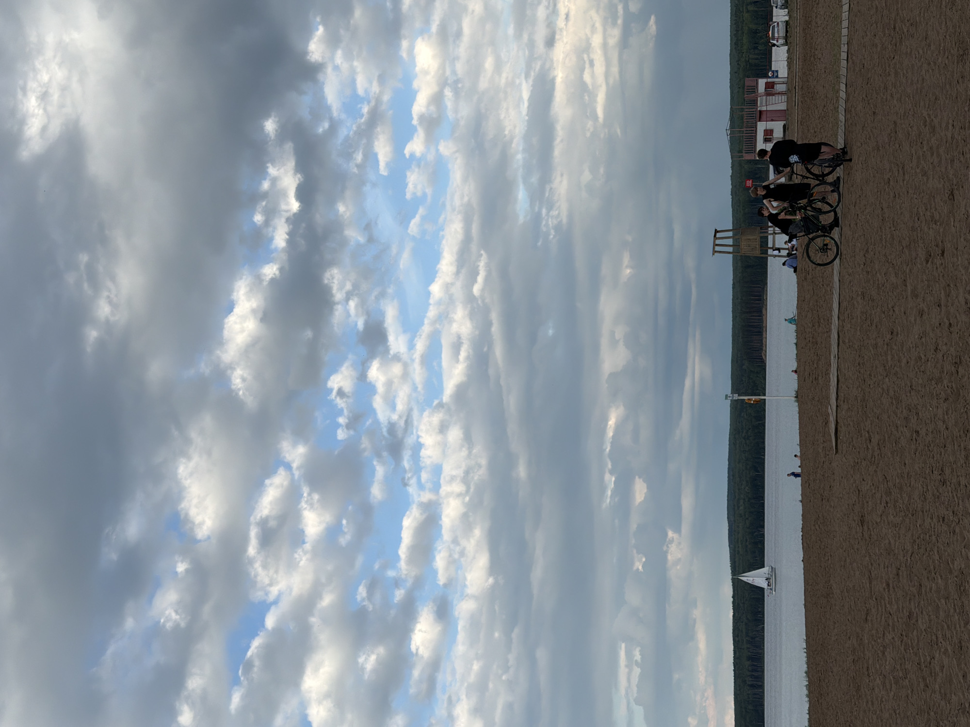 The image depicts a beach scene at the Kama River in Chaikovsky, Russia. In the foreground, three young men are gathered with bicycles on a sandy beach. They appear to be in casual conversation or resting. Further back, a body of water, likely the Kama River, is visible, with a sailboat on the horizon. Along the shoreline, there is a line of trees and various structures, including what appears to be a lifeguard tower and a small building. The sky is filled with dramatic, textured clouds, suggesting either an overcast day or the approach of weather. The overall atmosphere is one of a relaxed, late-afternoon or early-evening outing at a popular recreational area.
