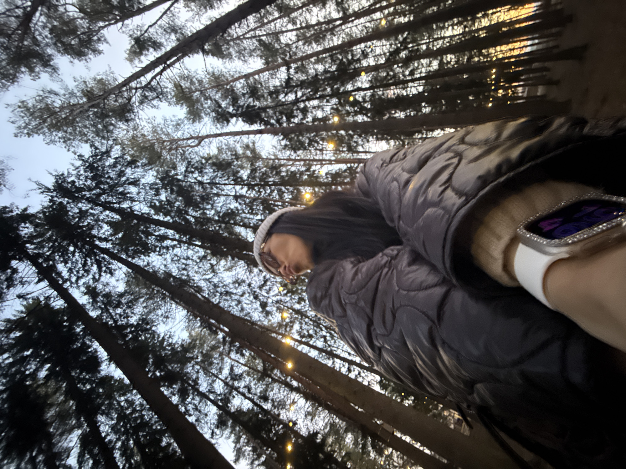 The image, taken in Votkinsk, Russia, captures a low-angle perspective of a person looking up at a canopy of tall trees adorned with fairy lights during dusk or early night.
The main subject is a person, visible from the torso upwards, positioned on the right side of the frame. They have long dark hair and wear a light-colored beanie, glasses, and a dark, possibly padded or puffer, jacket over a lighter long-sleeved garment. Their face is seen in profile, looking upwards and slightly to the right, suggesting they are observing the illuminated tree canopy. On their left wrist, a smartwatch with a white band is visible, featuring a screen with what appears to be a digital time display in purple on a dark background, surrounded by a bezel encrusted with sparkling elements.
The background is dominated by numerous tall, slender tree trunks and branches, silhouetted against a lighter, dusky sky that shifts from pale blue to grey. Strung horizontally between the tree branches are numerous small, warm-toned fairy lights, creating a soft, ethereal glow that punctuates the darkness. The illumination from these lights suggests the scene is set during twilight or after sunset.
The overall scene conveys a tranquil, contemplative mood, with the individual seemingly engrossed in the illuminated natural environment, possibly in a park or forest.