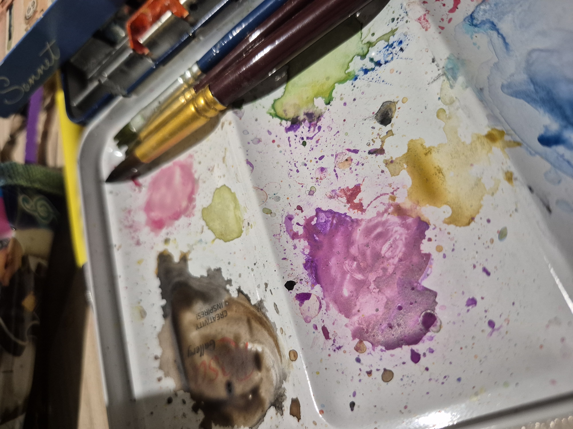The image shows a close-up, overhead view of a well-used watercolor paint palette. The palette is white and appears to be made of metal, with several wells containing dried and wet paint in various colors, including brown, pink, yellow, purple, green, and blue. Splatters and drips of paint are visible throughout the palette, indicating active use. Several paintbrushes are resting on the palette, with some appearing to be gold, brown, and blue. In the background, out of focus, is a dark blue tin case with the word "Sonnet" visible in cursive script. The scene is lit from above, casting subtle shadows. The image does not contain any recognizable landmarks or features that would suggest a specific location within Popovo, Bulgaria, or any other city.