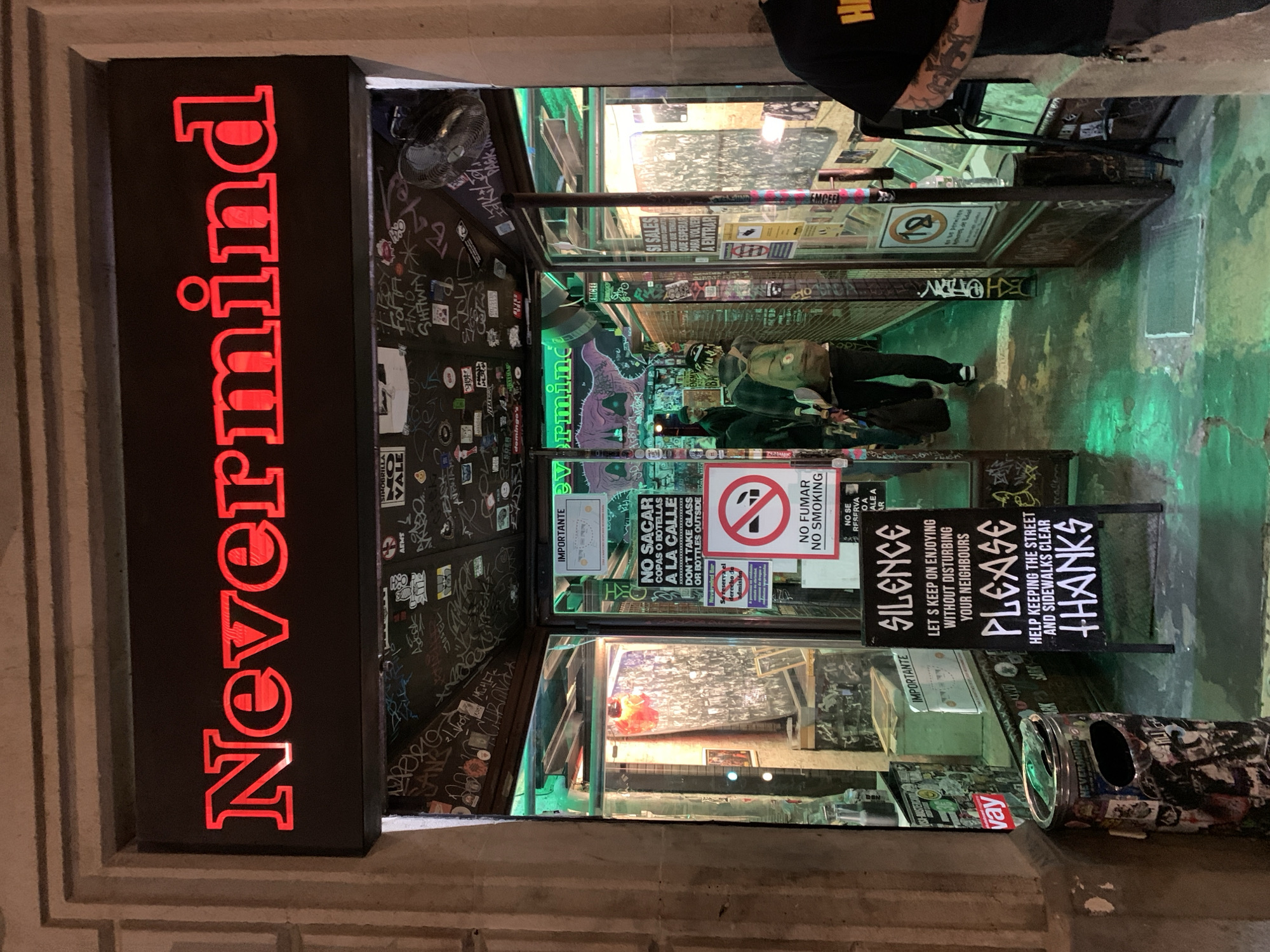 The image captures the entrance of "Nevermind," a bar or club in Barcelona, Spain, at night, characterized by its alternative and urban aesthetic. A prominent red neon sign vertically displays "Nevermind" on the left side of the frame. The entrance area is dimly lit with a distinctive green hue emanating from within, reflecting on the floor and glass surfaces.
Inside, two individuals are visible: one person with an olive green jacket and a backpack appears to be standing near the entrance, and another person with a partially visible black cap and tattooed arm is in the upper right. The walls and pillars around the entrance are heavily covered in graffiti, stickers, and worn textures, contributing to a grungy atmosphere. A black standing sign outside advises "SILENCE LET'S KEEP ON ENJOYING WITHOUT DISTURBING YOUR NEIGHBOURS" and "PLEASE HELP KEEPING THE STREET AND SIDEWALKS CLEAR THANKS." Another sign, partly obscured, reads "NO SACAR COPAS O BOTELLAS A LA CALLE DON'T TAKE GLASS OR BOTTLES OUTSIDE" and "NO FUMAR NO SMOKING." A "No 18" sign (likely indicating an age restriction) is also visible. A sticker-covered trash can with a metallic lid stands near the right side of the entrance. The overall scene suggests a vibrant, late-night venue with an edgy, counter-culture vibe.