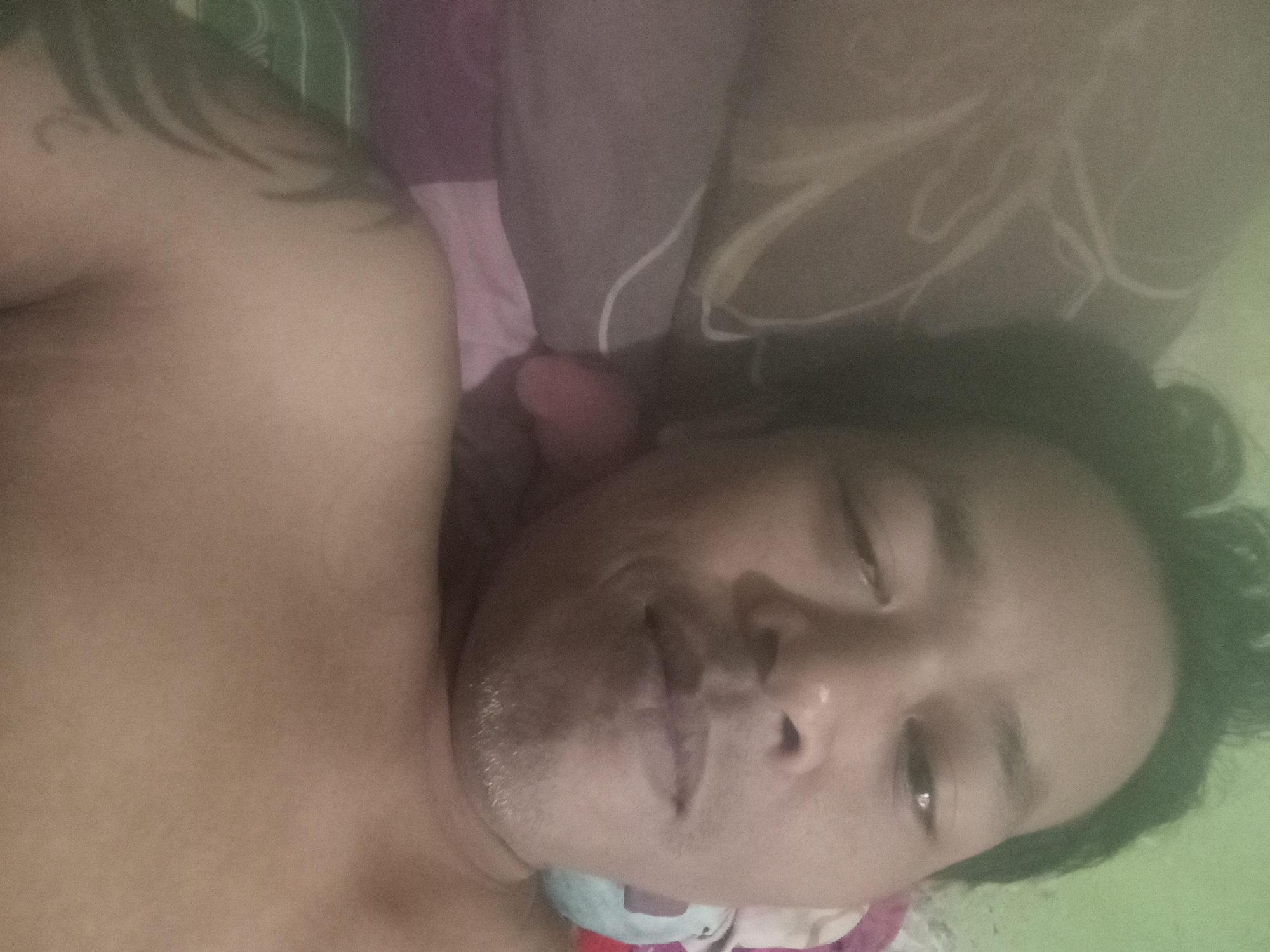 A selfie is taken by a male person lying down on a bed. The person has dark hair and fair skin. The person is shirtless and has a tattoo on their left shoulder. The background shows pillows and a green wall. The image is blurry and poorly lit.