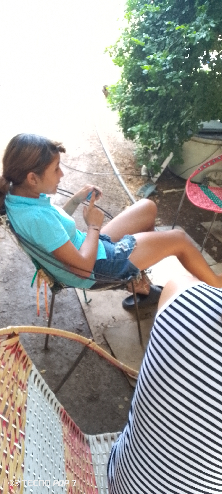 A young woman with brown hair tied back is seated in profile in a black and green canvas chair, actively engaged with a smartphone held in both hands. She is wearing a light blue polo shirt and distressed denim shorts. Her right index finger is extended towards the phone's screen, suggesting interaction. A black flip-flop rests on the ground near her feet.
The setting is outdoors, likely a shaded patio or backyard in Altagracia, Venezuela. The ground is composed of dirt or gravel, with a light-colored pipe or hose running across it. Lush green foliage and bushes are prominent in the background on the right side of the image, contributing to the natural outdoor ambiance. Another red and green woven chair is visible in the mid-ground to the right. In the immediate foreground, a portion of a red and white woven chair is visible, and behind it, a person wearing a black and white striped top is partially seen.
The bright, natural lighting indicates it is daytime. The overall mood appears relaxed and casual, with the main subject focused on her device. The watermark "TECNO POP 7" is visible in the bottom-left corner of the image.