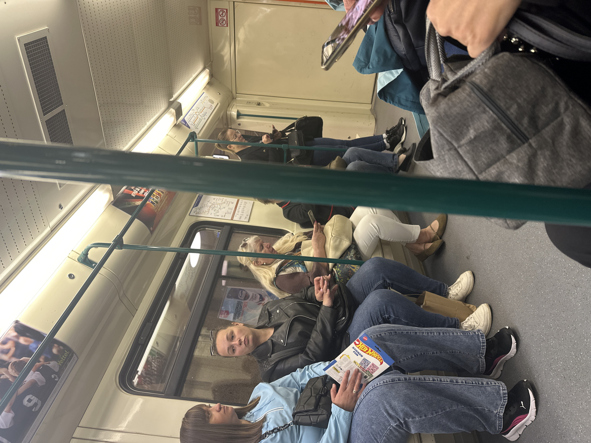 Inside a subway car, presumably in Sofia, Bulgaria, judging by the Cyrillic signage. Several women are seated, some occupied with phones or books. The car's interior appears utilitarian, with off-white walls and metal handrails. Passengers are dressed in casual attire, including jeans, jackets, and sneakers. Some personal belongings, like bags, are visible.