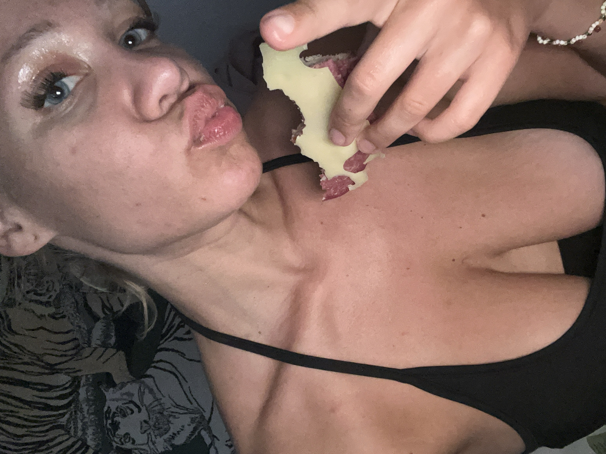 A young woman with blue eyes and blonde hair is posing for a selfie. She is wearing a black top and has a piece of cheese and cured meat in her hand. The background shows a tiger-patterned tapestry. The image was taken in Mühldorf a. Inn, Germany.