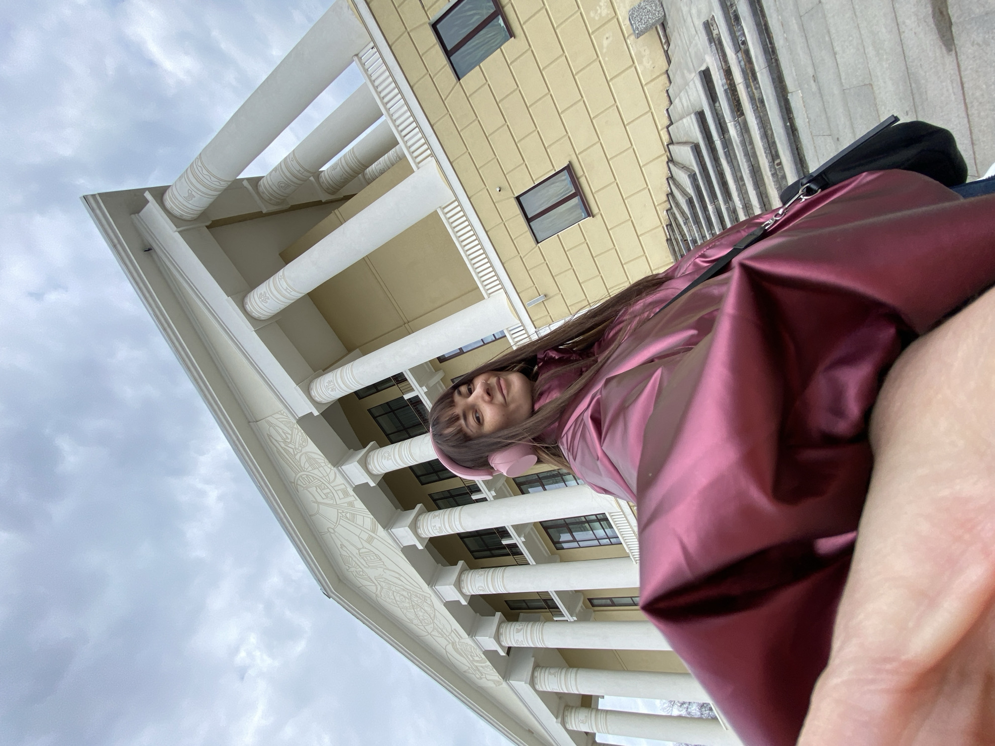 A low-angle shot depicts a woman wearing headphones and a shiny burgundy jacket, positioned in front of a building in Votkinsk, Russia. The building features white pillars and a decorative facade. Windows are visible on the structure behind the pillars. A grey sky is overhead, and stairs are located to the right of the building. The woman appears to be taking a selfie.
