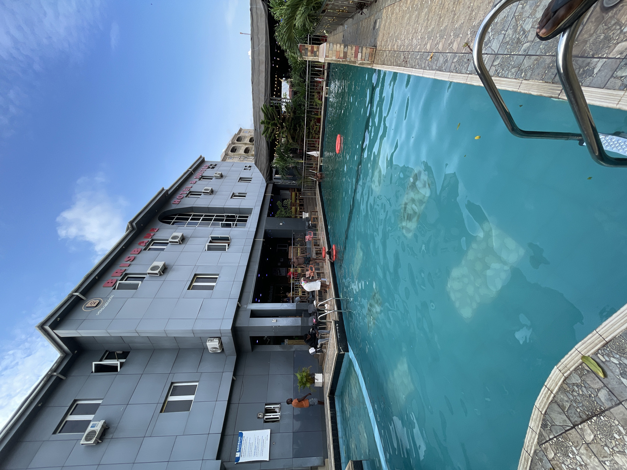 A hotel and swimming pool scene is captured in this tilted, eye-level medium shot. The primary subjects are the Brisbane Hotel & Suites building, the swimming pool, and several people.
The building, a multi-story structure with a grey, tiled facade, features numerous windows and air conditioning units. Text visible on the building reads "BRISBANE HOTEL & SUITES." A logo with the letters "MM" is also present. To the right of the building is a vibrant blue swimming pool with a stainless-steel ladder. Reflections of the building and surrounding trees are visible on the water's surface.
Several individuals are scattered around the scene. One person is partially submerged in the pool, and another sits on the edge of the pool. Others are seated at tables and chairs near the pool, suggesting a relaxed atmosphere. An orange flotation ring is floating in the pool. The setting appears to be a resort or hotel complex, likely in a tropical climate given the presence of palm trees.
The time of day appears to be afternoon, with bright daylight and a clear blue sky. The weather seems pleasant, suitable for swimming.
The overall impression is one of leisure and relaxation, with a focus on the amenities provided by the hotel. The presence of both the hotel building and the pool indicates a commercial establishment catering to guests. The image is taken from a slightly elevated perspective, looking down towards the pool and up towards the building.