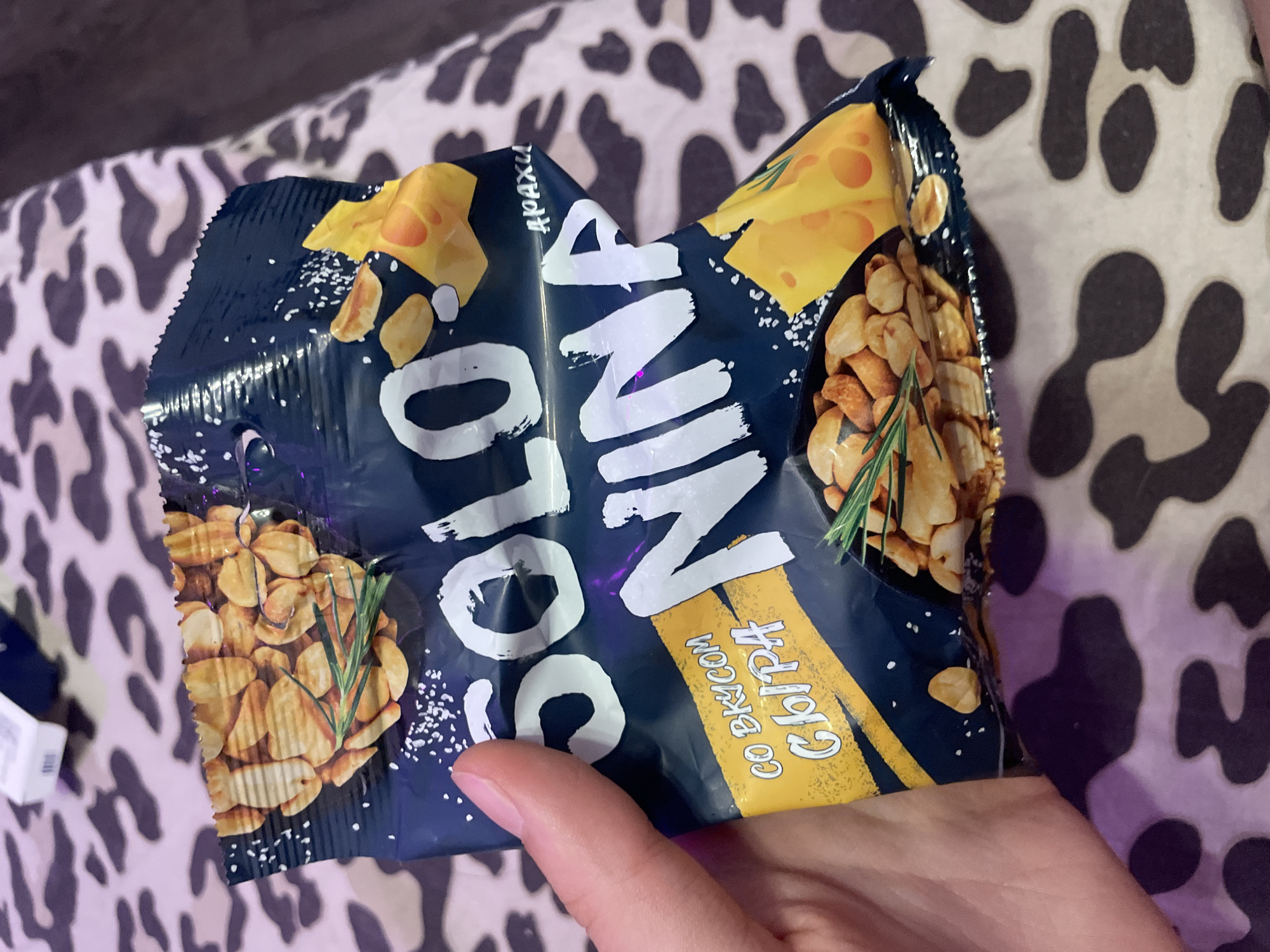 A hand is holding a crumpled dark blue snack bag with images of peanuts, cheese, and rosemary. The bag has white text that reads "SOLO NINA" and "СО ВКУСОМ СЫРА" (with cheese flavor). The background is a patterned blanket or fabric. The image appears to be taken indoors. There is no information about Vaganovo, Russia in the image.