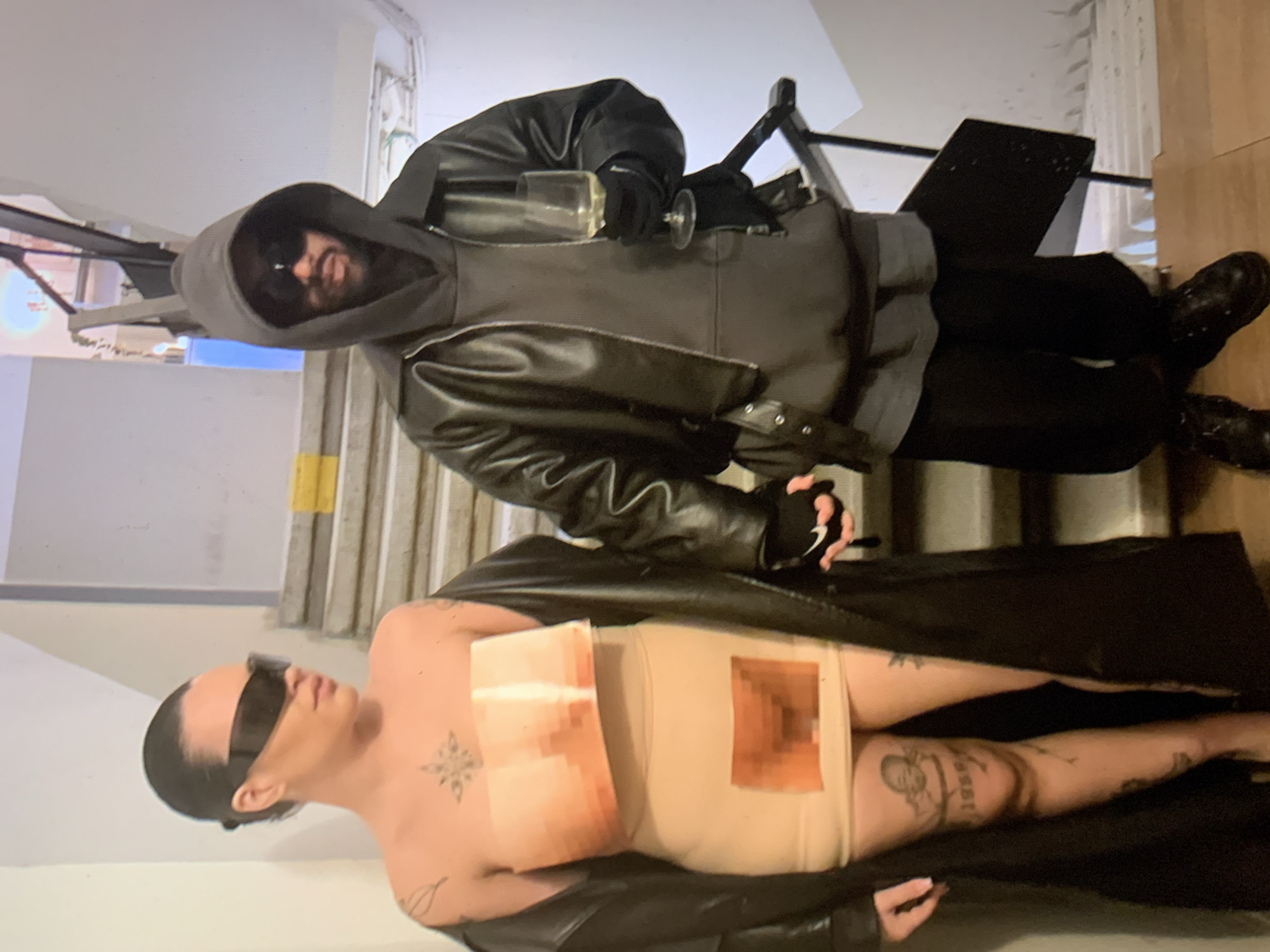 The image appears to depict two individuals on a staircase. The individual on the upper portion of the frame is wearing a dark hoodie, black leather jacket, black gloves, and sunglasses and is holding a glass of wine. The person on the lower portion of the frame is wearing a nude body covering, sunglasses, and a long dark coat. The person on the lower portion of the frame also appears to have tattoos and has pixelated covering on their body covering. The location appears to be indoors on a staircase. There is no indication that the image was taken in Tel-Aviv, Israel.