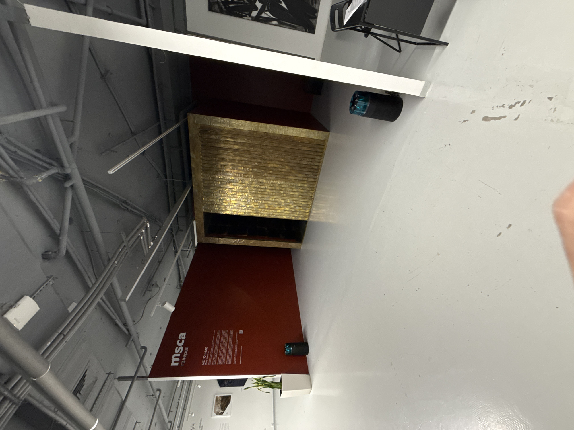The image shows an interior view of the MSCA (Moscow Contemporary Art) gallery in Moscow, Russia. The camera angle is off-center, and the image has been rotated.
On the left side, exposed grey piping and structural elements of the building are visible on the ceiling. A red wall with the "MSCA" logo and Russian text is present, with a small green plant in a white rectangular pot at the bottom.
Above the red wall, there's a golden, textured structure with a dark opening underneath. Above this is another, deeper red section, topped by a white horizontal structure. In the upper part of the image, a black-and-white framed artwork hangs on the wall above a dark structure.