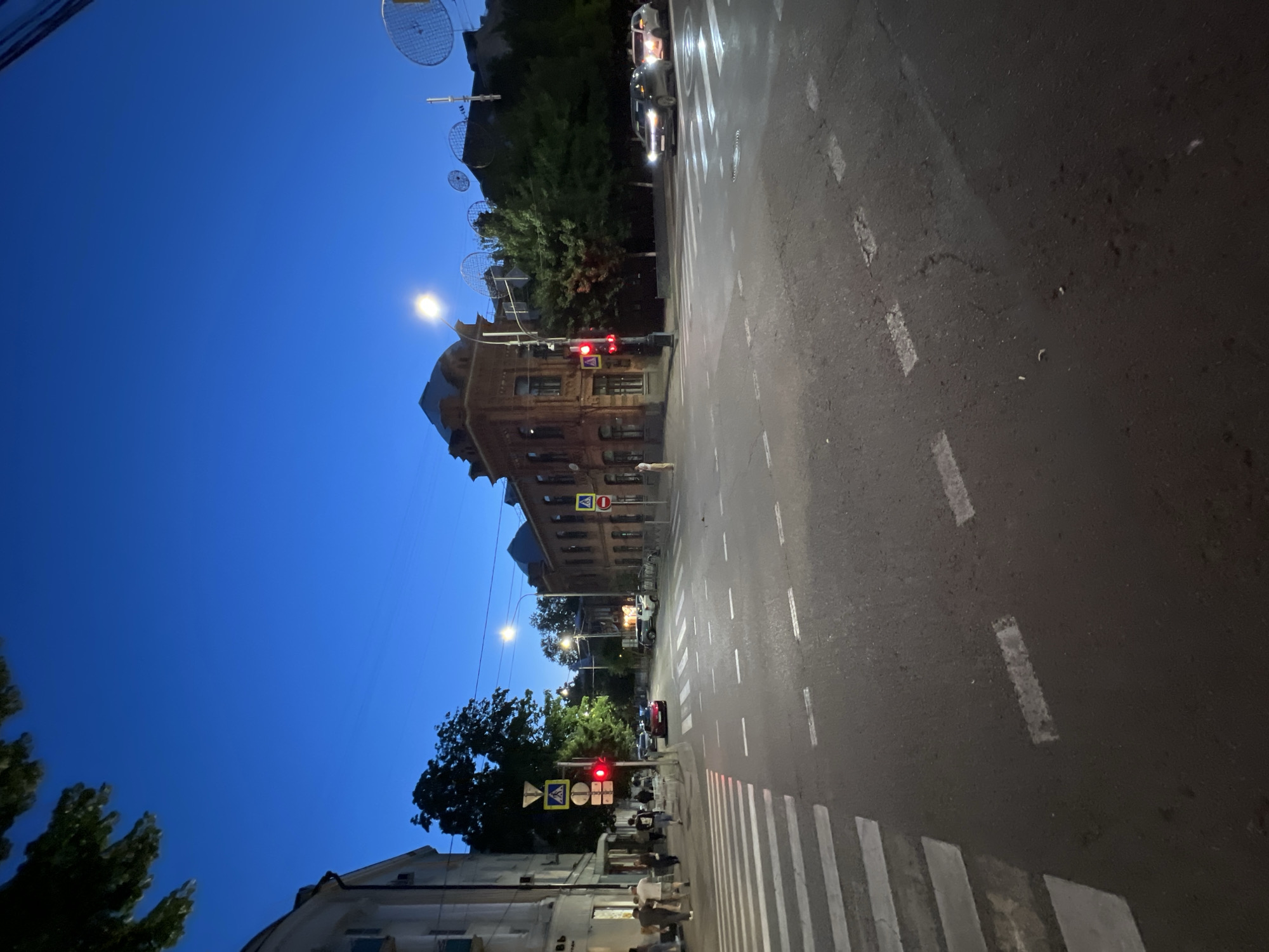 This media file depicts an outdoor scene at dusk in Krasnodar, Russia. The image is oriented vertically, giving it a unique perspective of a street and adjacent buildings. The sky is a deep, clear blue, indicative of the twilight hour.
The street features a crosswalk with prominent white stripes. Lane markings are visible on the asphalt, suggesting it's a moderately busy road. A traffic light is illuminated with a red signal, indicating a stop. Several cars are present, with headlights on, implying low visibility due to the time of day.
Buildings line the street, one of which is a multi-story brick structure with architectural details suggesting it's an older building. Trees with green foliage are interspersed along the street, some appearing quite tall and lush. Streetlights are also visible, casting a warm glow.
Several people are depicted near the crosswalk and sidewalk. Their presence suggests daily urban life is continuing as evening sets in. The overall mood is calm and ordinary, capturing a typical moment in an urban environment transitioning from day to night. There are no discernable emotional expressions captured in the individuals.
Visible signs include road signs with red circles and white bars, likely indicating traffic restrictions or prohibitions, and yellow diamond-shaped signs with black symbols, common for warnings or directions. There is no legible text on the buildings or vehicles. The weather appears clear and calm.