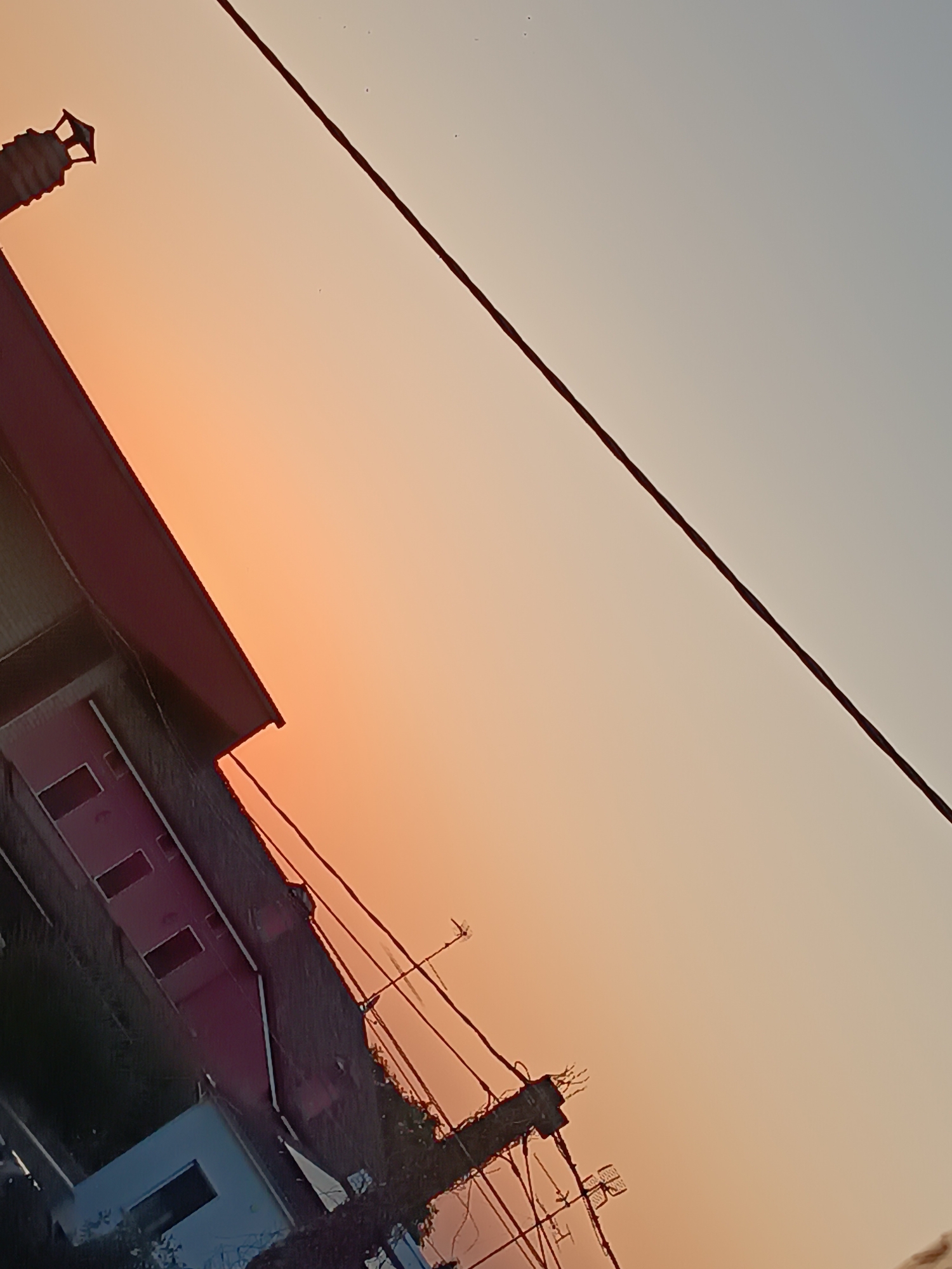 A low-angle outdoor shot captures a building with a red roof and dark walls, possibly brick, featuring several rectangular windows. The building is partially obscured by a power line that diagonally bisects the frame from the top right to the bottom left. The sky exhibits a gradient of sunset colors, transitioning from a warm orange near the horizon to a pale blue overhead. Several small, dark dots are visible in the sky, potentially birds or dust motes. A lamppost-like structure with a geometric cap is visible on the left side of the frame, near the top of the building. In the lower part of the image, more power lines and poles are apparent, along with some foliage. The scene suggests an urban or suburban environment during twilight.