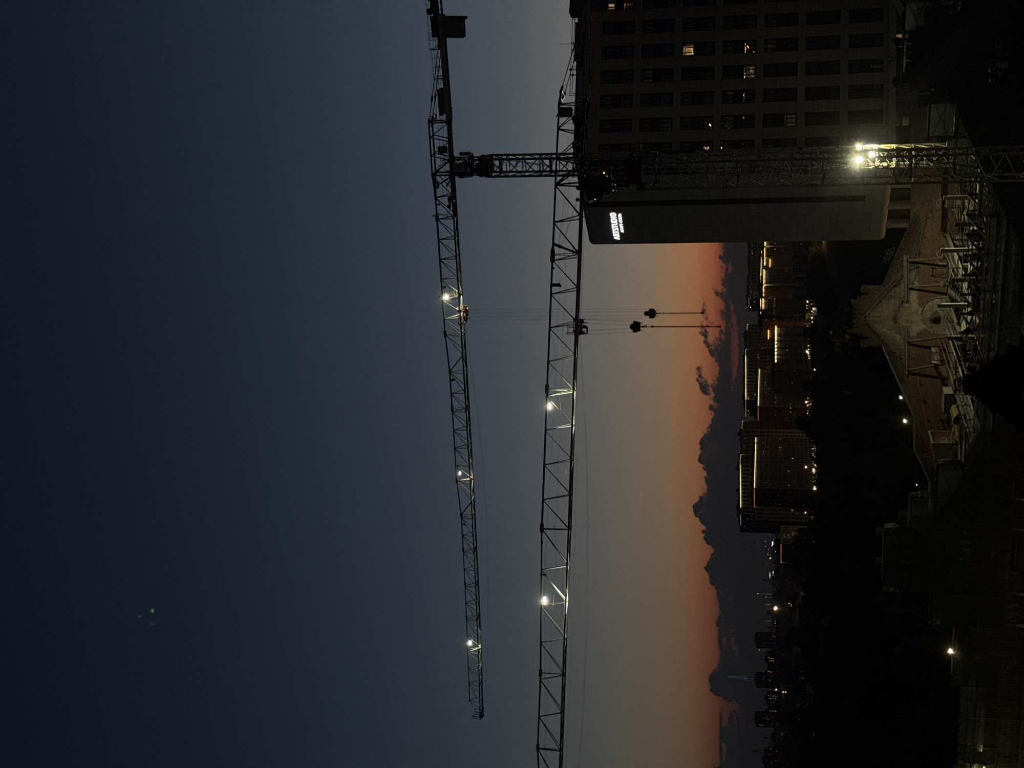 The image depicts a twilight scene in Sinyavino, Russia, with construction cranes dominating the skyline. Two large tower cranes are prominently featured, their metallic structures silhouetted against a gradient of deep blue to orange sky. Lights are visible along the jib of each crane, illuminating their long beams. In the mid-ground, a modern building with illuminated windows and the "ARTSTUDIO" logo is visible, adjacent to one of the cranes. Further in the background, a cityscape is discernible with more buildings featuring illuminated facades and lights. Dark clouds are scattered across the horizon, adding depth to the dusk sky. The foreground is mostly dark, obscuring finer details of the ground level, but suggests ongoing construction activity with some visible structures and lights. No people are discernible in the image.