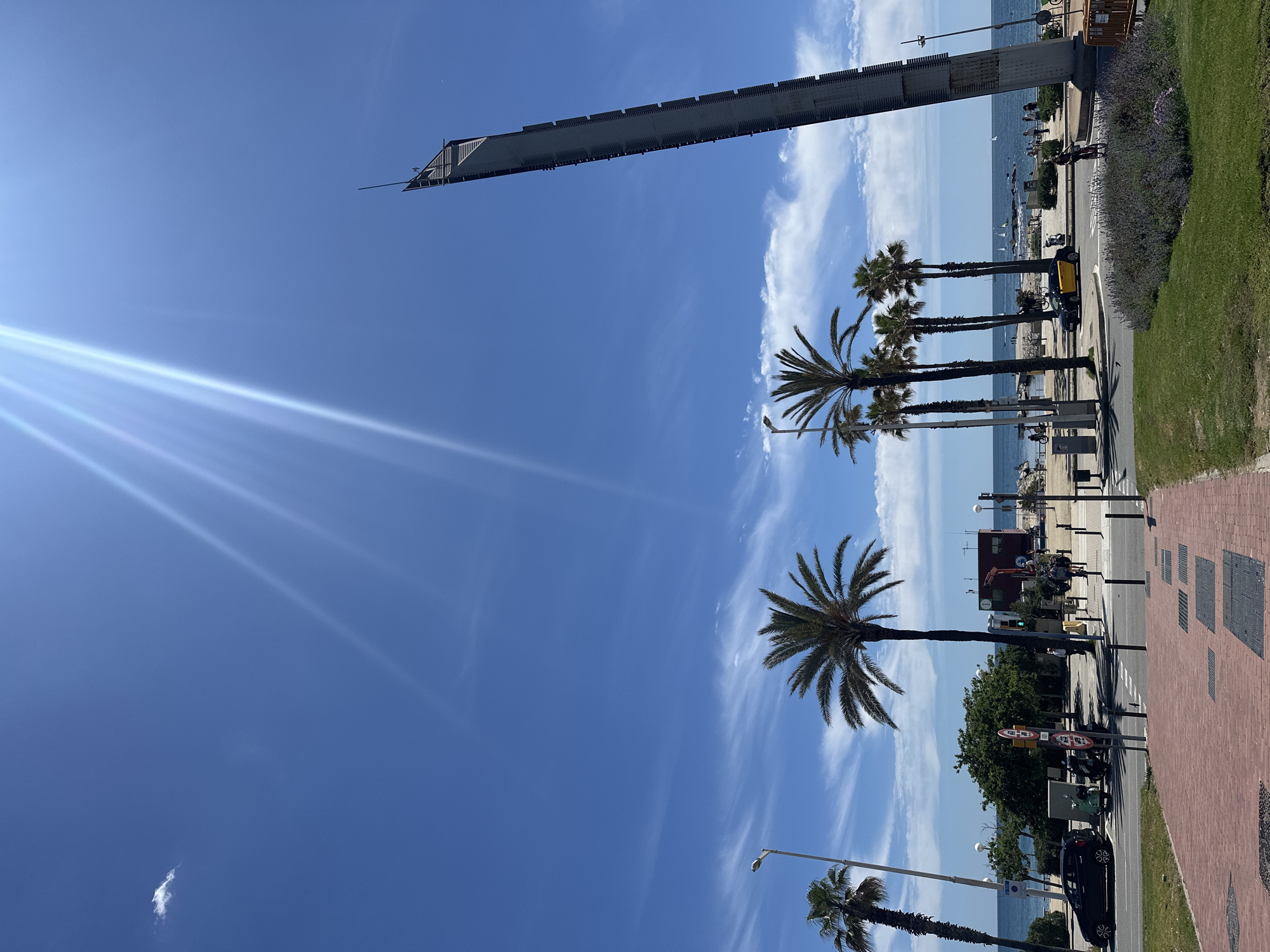 The image captures a street scene in Barcelona, Spain, featuring palm trees lining the road adjacent to a beach. A notable architectural structure, the Estela de Barcelona, dominates the upper part of the frame. The sky is clear blue with a few streaks of clouds and visible sun glare. Vehicles and pedestrians are present along the road and on the beach, indicating a typical urban-beach environment. A brick sidewalk runs along the edge of the road. The overall atmosphere is sunny and bright.