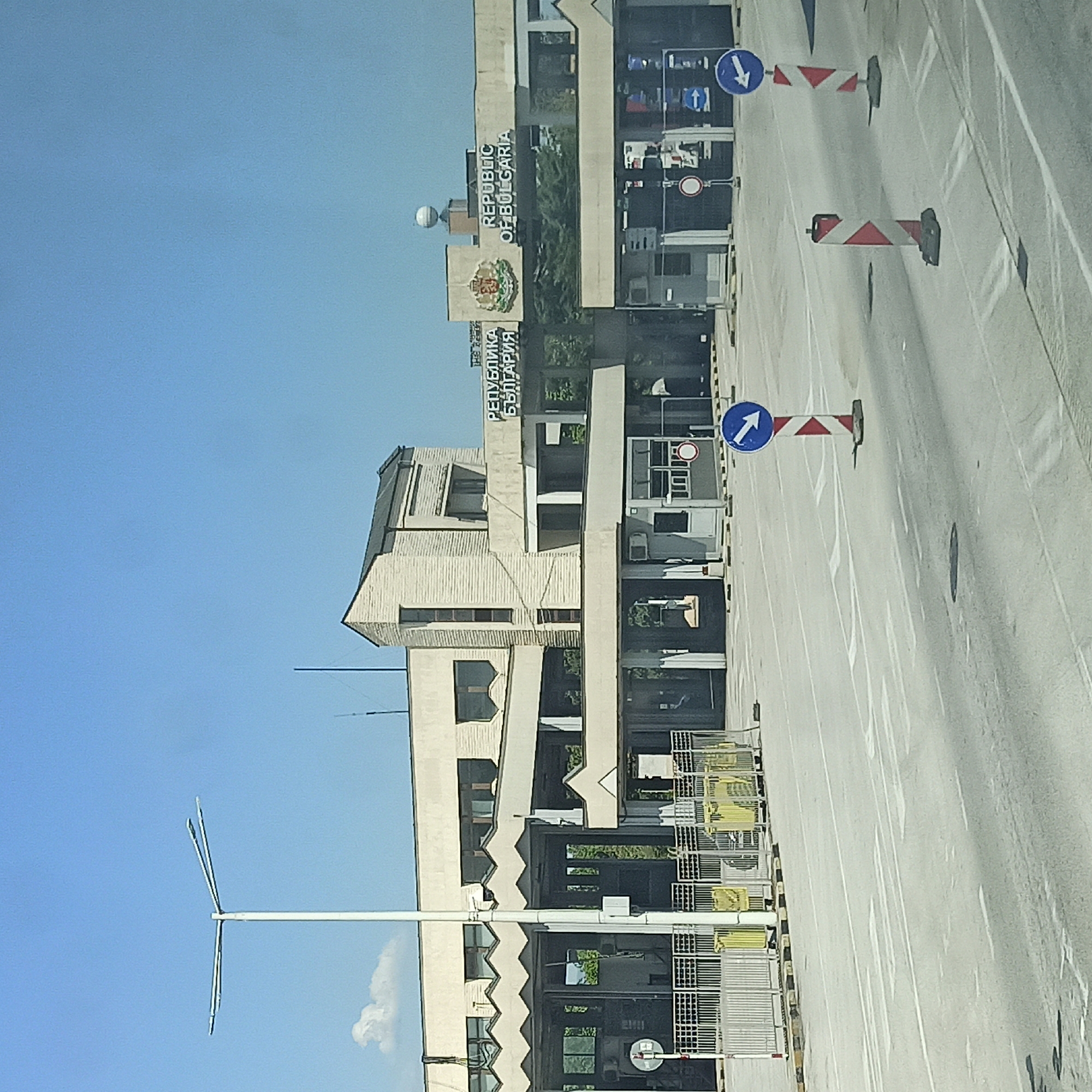 The image shows the Kulata border crossing between Bulgaria and Greece. The scene depicts a border control area with several buildings, including a customs house with the words "REPUBLIKA BALGARIYA" and "REPUBLIC OF BULGARIA" visible on its facade. The buildings are modern, constructed with light-colored materials and large windows. Various traffic signs, including directional arrows and regulatory signs, are placed throughout the area. Road markings are visible on the asphalt surface. Security fences and barriers are present, along with some yellow structures that might be queuing lanes or bollards. The sky is clear and blue, indicating daytime. There are no people visible in the immediate foreground or prominent in the scene, suggesting a quiet period at the border crossing. The overall impression is a functional and orderly border control facility.