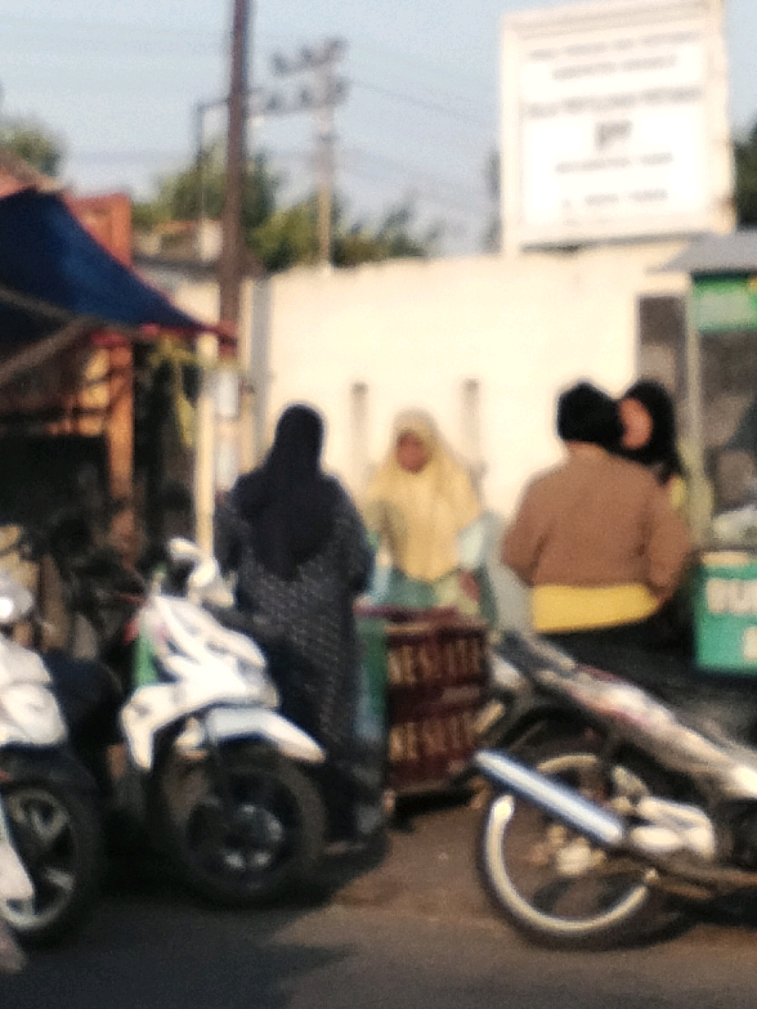 The image depicts a street scene in Singogalih, Indonesia, likely a market or roadside stall area. In the foreground, two motorcycles are parked, one on the left with its front wheel visible and a white and black body, and another on the right, partially obscured, with a silver exhaust pipe and black wheels. The motorcycle on the right appears to have a wooden crate or basket behind it.
Several people are present. On the left, a person wearing a black hijab and a patterned dress is standing, their back to the camera. To their right, a person in a light yellow hijab is also visible. Further to the right, two individuals are standing near each other. One is wearing a brown jacket over a yellow shirt, and the other person's face is partially visible, wearing a dark top.
The background includes a white building with some signage, though the text is blurry. There are also power lines and trees visible, suggesting an outdoor urban or semi-urban environment. The overall lighting indicates it could be daytime, possibly late afternoon given the softness of the light. The image is slightly out of focus, adding a soft, almost impressionistic quality to the scene.