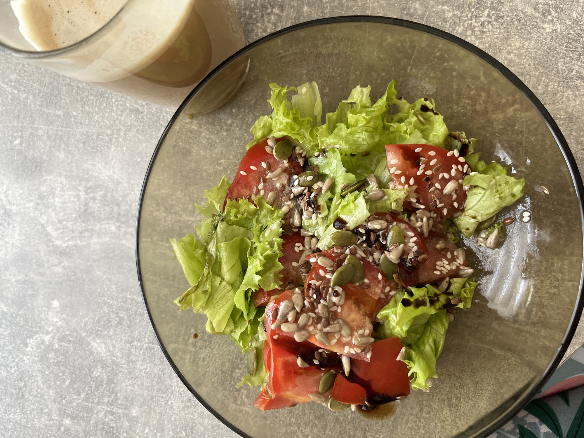 A top-down, high-angle shot captures a glass bowl filled with a salad composed of lettuce, sliced tomatoes, and various seeds, dressed with a dark sauce. A portion of a glass containing a frothy, layered beverage is visible in the upper left corner of the frame. The salad sits on a gray surface with subtle variations in texture. The setting is Chaikovsky, Russia.