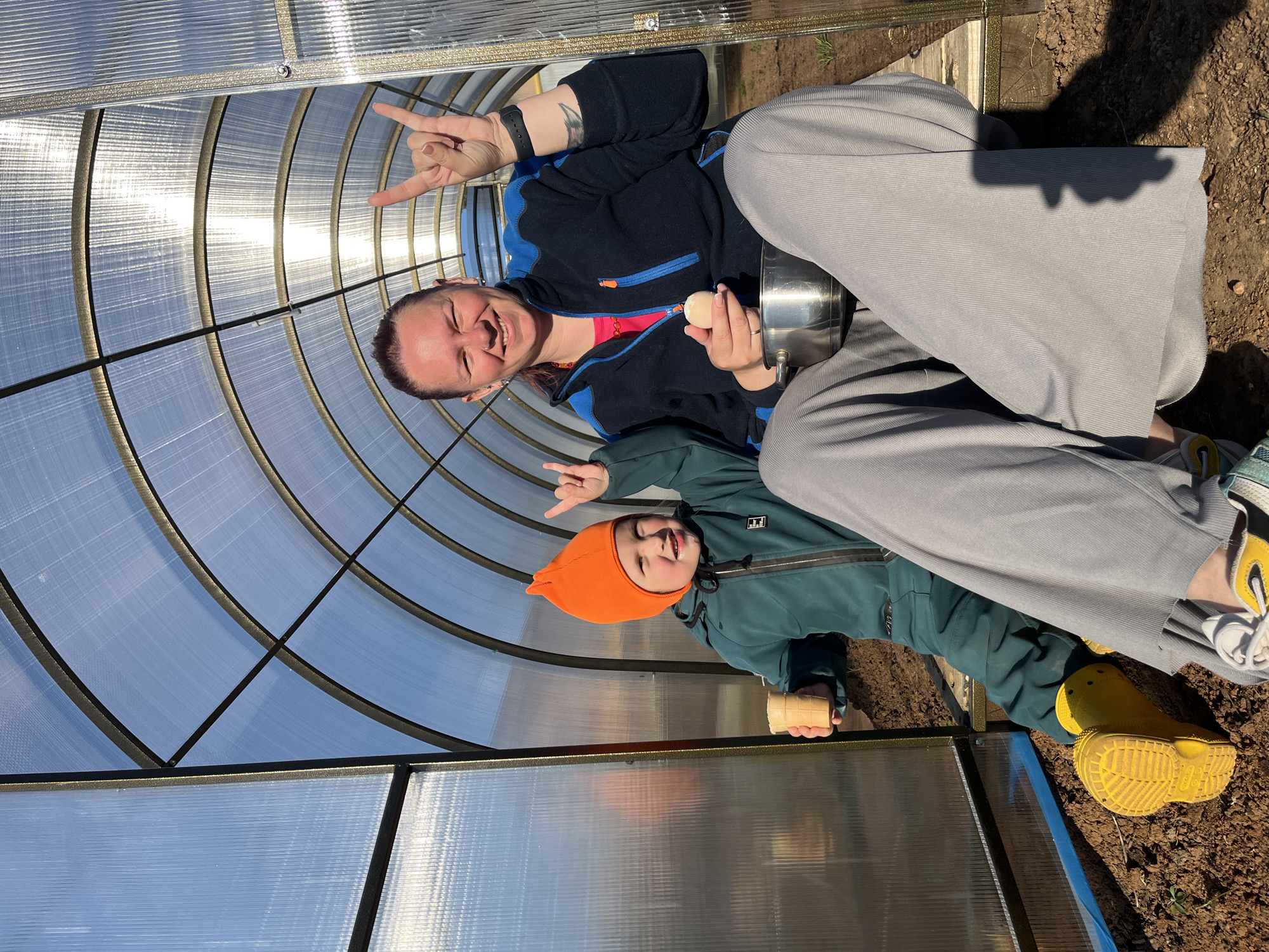 The image shows two people, likely a mother and child, inside a greenhouse in Chaikovsky, Russia. The mother is sitting and holding a pot in one hand and an egg in the other. She is making a "peace sign" with her left hand. She is wearing a dark blue jacket with blue accents, a pink shirt underneath, and light grey pants. The child, wearing a green jacket and orange hat, is also making a "peace sign" with their hand. The greenhouse is made of transparent corrugated plastic panels, and the ground outside is brown soil. The child is wearing yellow boots.