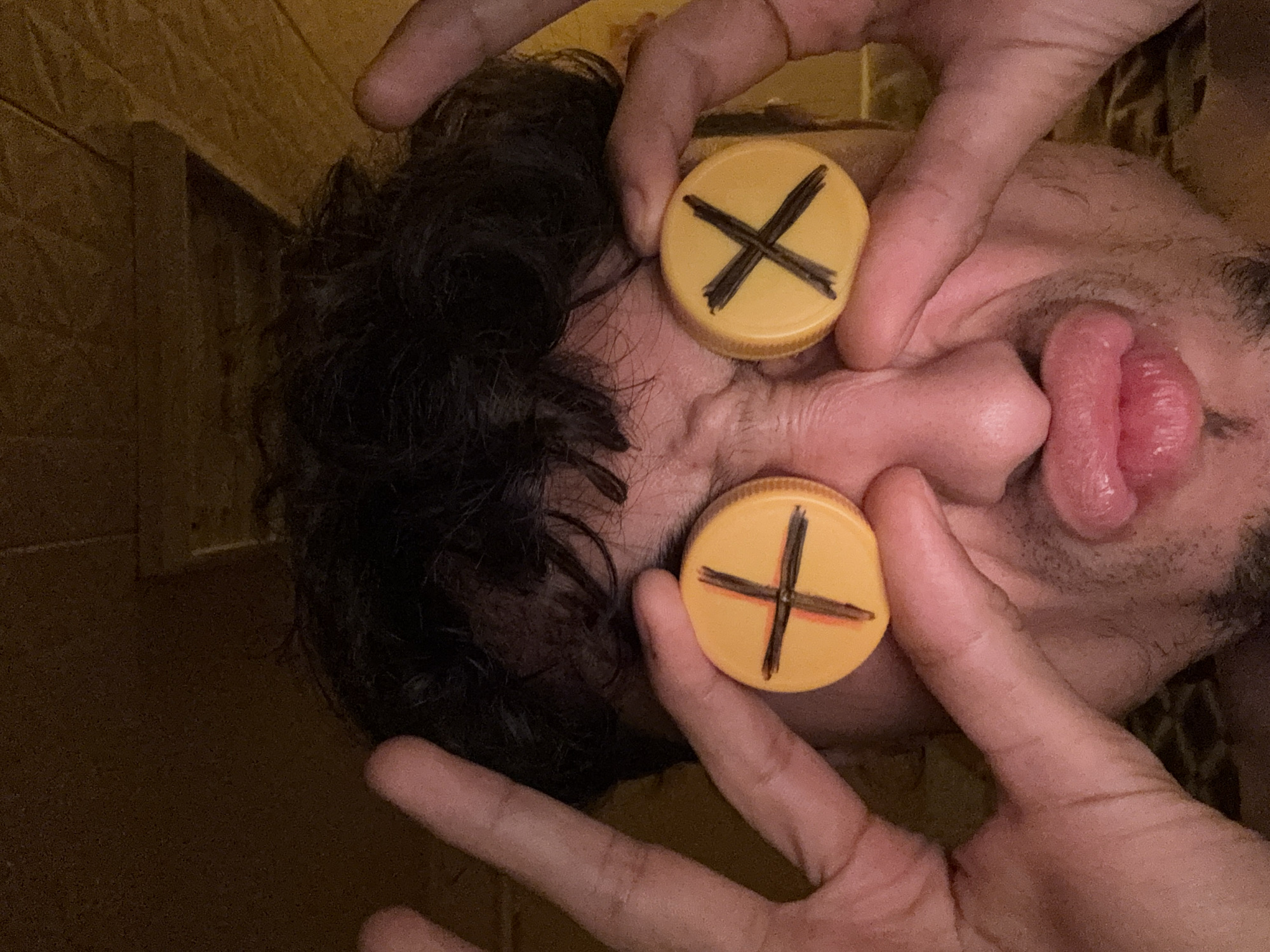 The media file shows a close-up, head-on shot of a person's face, viewed from a slightly upward angle. Two bottle caps, colored yellow and marked with black "X" shapes, are held over the person's eyes by their hands. The person's mouth is open, with their tongue slightly visible, and their lips are pink and appear moist. The background is a textured, brown surface, possibly a wall, with some darker areas suggesting shadows or uneven lighting. The overall lighting appears to be warm and somewhat dim, creating a moody atmosphere. The focus is sharp on the bottle caps and the person's face, with the background being slightly out of focus. The hands holding the bottle caps are clearly visible, with fingers extended. There is no visible text in the image. The image has a playful, perhaps even humorous, or staged aesthetic due to the placement of the bottle caps over the eyes.