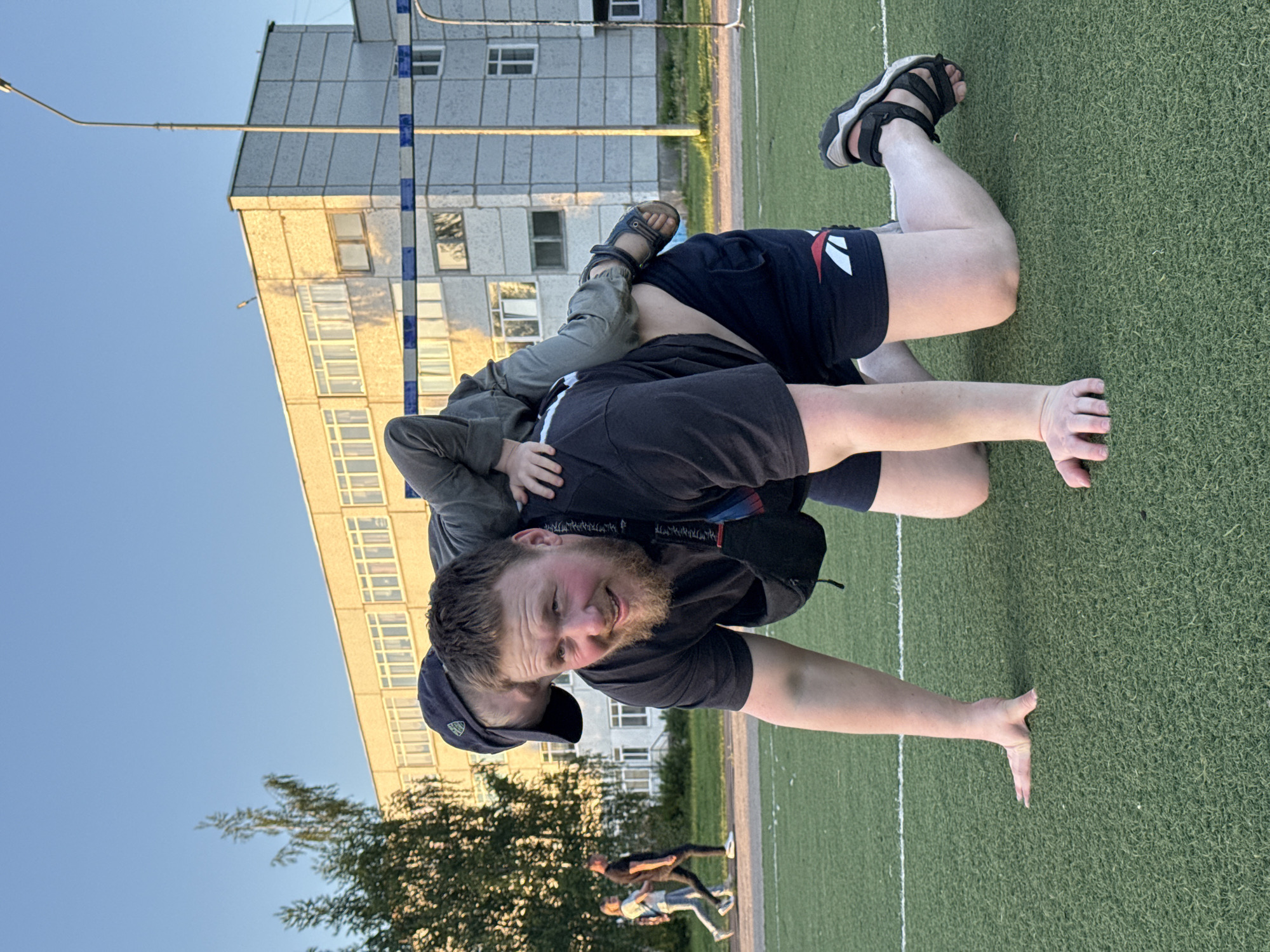 A man is on his hands and knees on an artificial turf field with a child on his back. The man is wearing a black t-shirt, dark shorts, and black sandals. The child is wearing a grey jacket and dark sandals. In the background, there is a light yellow building and a clear blue sky. Three people can be seen running in the background. There is a tree in the lower left corner. The location is in the city of Chaikovsky, Russia.