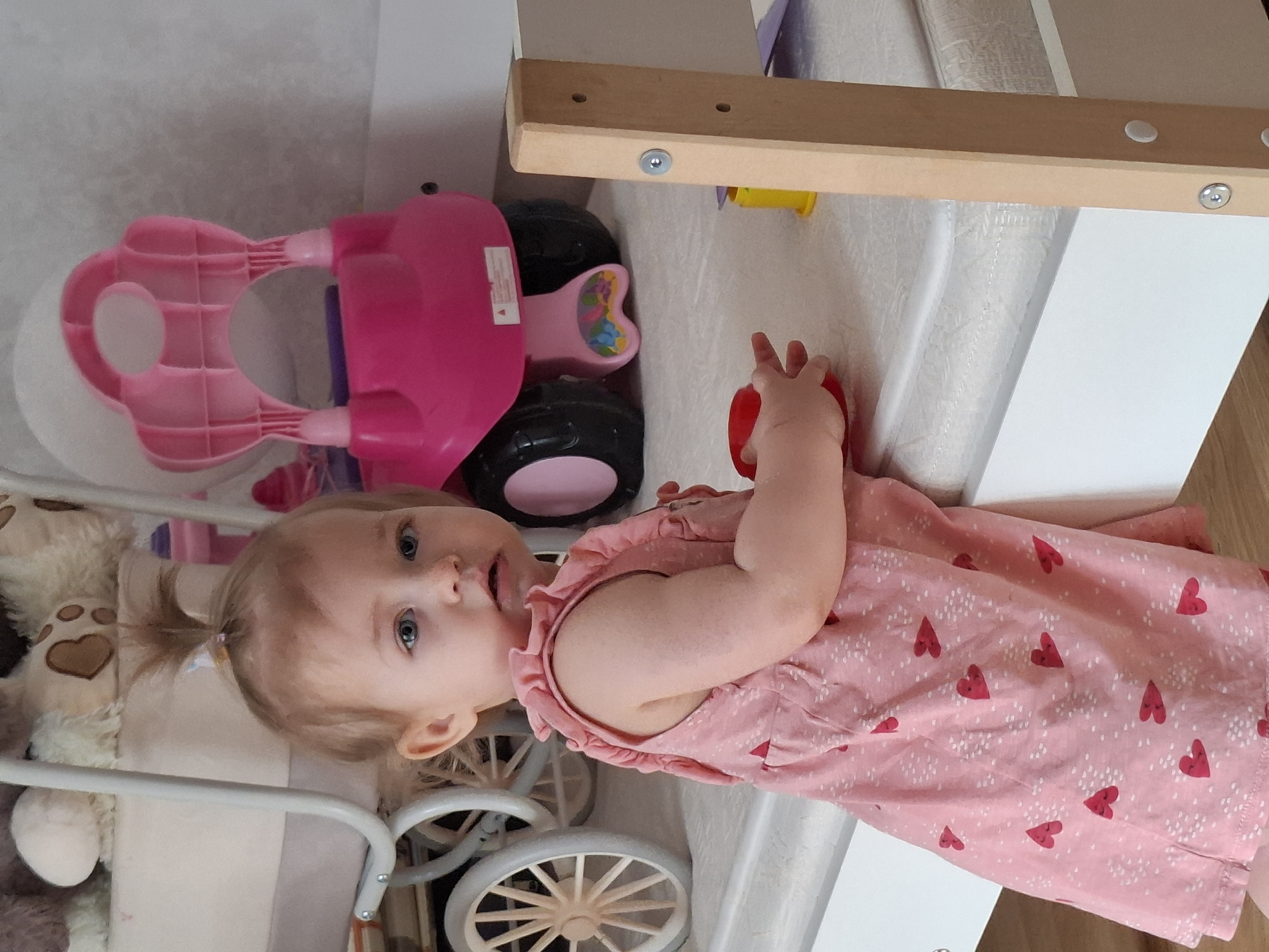 This media file features a light-haired toddler with blue eyes, wearing a sleeveless pink dress adorned with red hearts and white speckles. Her light-colored hair is styled in a small ponytail. The child is looking directly at the camera with an open-mouthed, curious expression, while holding a red object in her right hand.
She is situated indoors, appearing to be on a white mattress or soft surface, positioned beside a white structure with wooden beams, possibly part of a bed frame, crib, or playpen. To the child's left, a large pink plastic ride-on toy with black wheels is visible. Below her, partially visible, is a white toy stroller or pram with intricate, light-colored wooden spoked wheels. A tan stuffed teddy bear with embroidered brown heart-shaped paw prints can be seen near the stroller. The setting is brightly lit, suggesting daytime, within a domestic environment, likely a child's room or play area. There is no clearly legible text visible in the image. The location context for this scene is Chaikovsky, Russia.