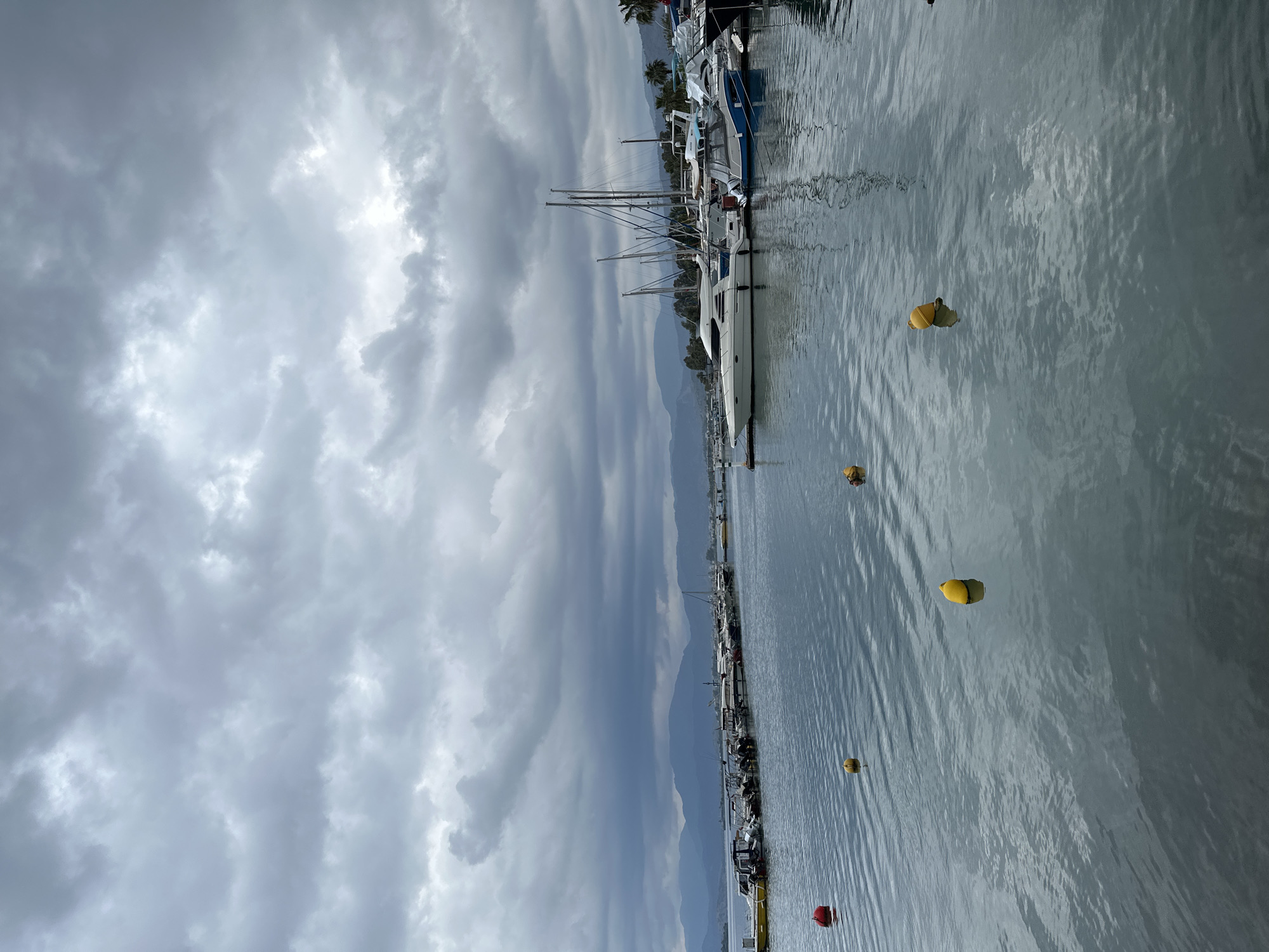This photograph captures a harbor scene, likely within the city of Fethiye, Turkey. The image is dominated by a cloudy, overcast sky. The water in the harbor is gray and slightly rippled, with several yellow and one red buoys floating on the surface. A row of boats, including sailboats and other smaller vessels, are docked along the harbor's edge. In the background, a hazy mountain range is visible.