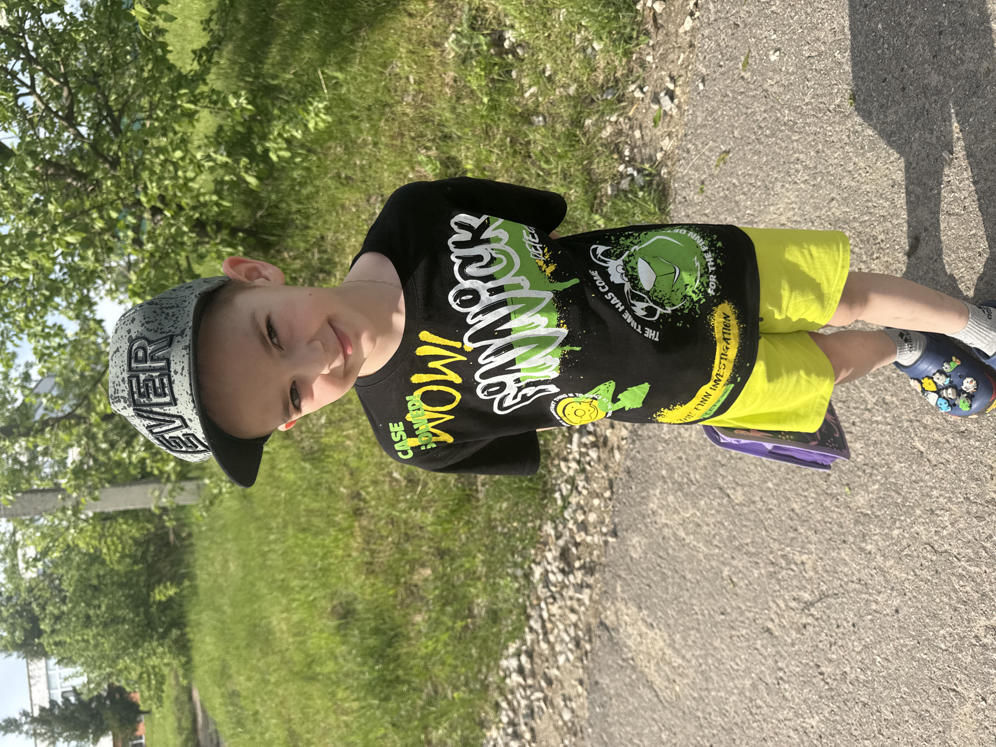 A young boy stands on a paved surface, likely a sidewalk or road, next to a grassy area with trees. He wears a black t-shirt with a graphic design featuring cartoon-like elements and yellow text, bright yellow shorts, and navy blue Crocs with character decorations. He also wears a gray and white patterned baseball cap. The boy is holding what appears to be a purple book or folder. The location may be Shevlyakovo, Russia.