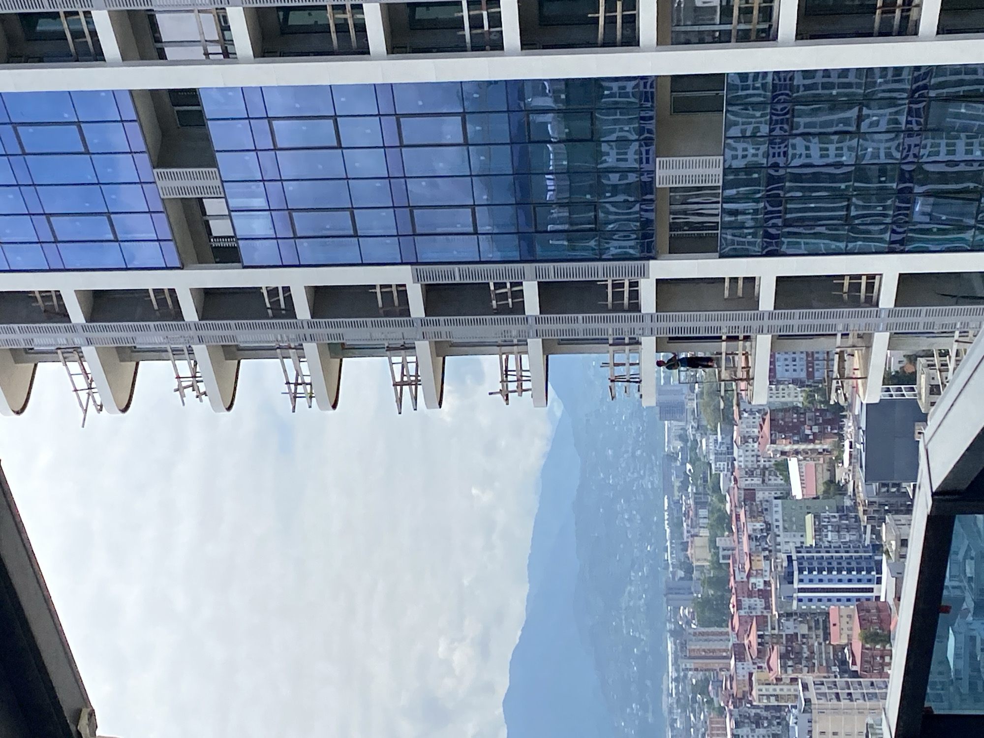This is a medium-angle, eye-level shot of a residential building under construction in Batumi, Georgia. A single construction worker, wearing dark clothing, is visible on a balcony of the building, standing near a railing. The building features a facade of blue-tinted glass windows and white concrete balconies with unfinished wooden railings. In the background, a dense cityscape with numerous residential buildings of varying heights and architectural styles is visible, with rolling, blue-hued mountains in the far distance under an overcast sky.