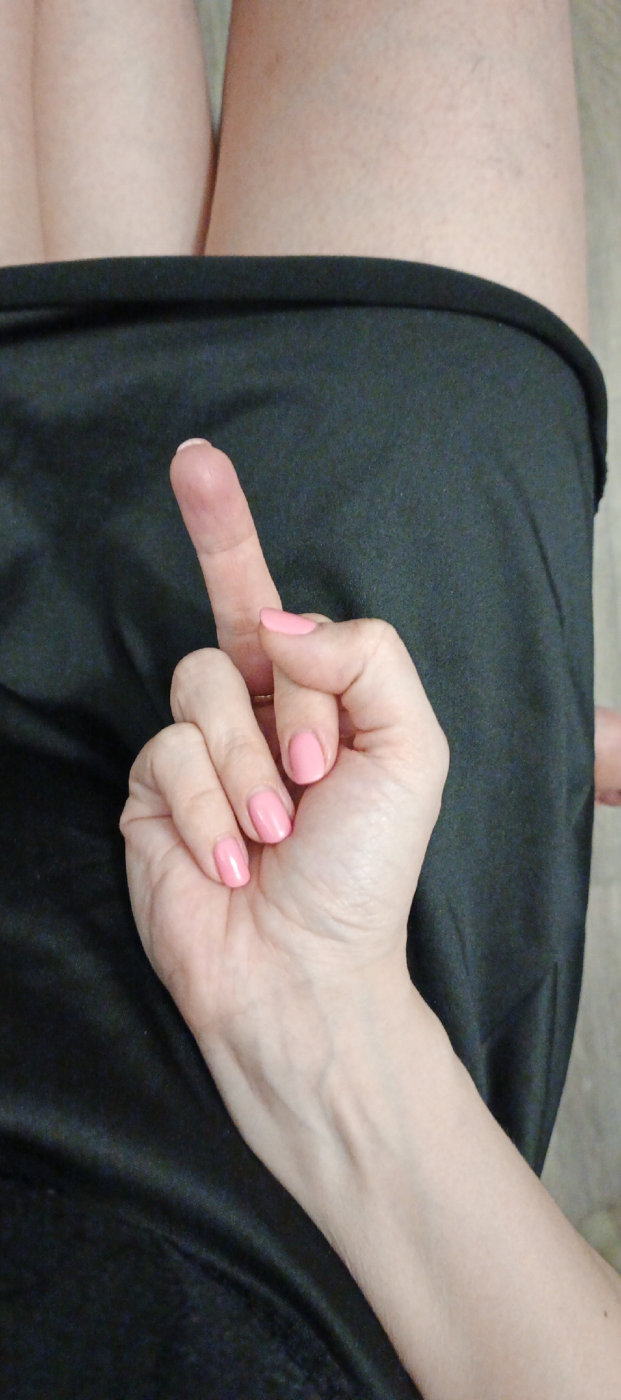 The image presents a close-up view of a person's hand, centered on an offensive gesture. The hand has a fair skin tone, and the fingernails are painted with a vibrant pink polish. The middle finger is extended straight upwards, while the other fingers are curled into the palm, emphasizing the gesture. A slender ring is visible on the ring finger.
In the background, a dark, presumably black, piece of fabric, likely clothing such as shorts or a skirt, partially covers the upper thighs of a person. Parts of the person's bare legs are visible above the dark fabric, suggesting a casual indoor setting. The specific time of day or weather conditions are not discernible from the image. The gesture clearly conveys emotions of defiance, anger, or contempt. No text is visible within the image. The provided location context for this media file is Chaikovsky, Russia.