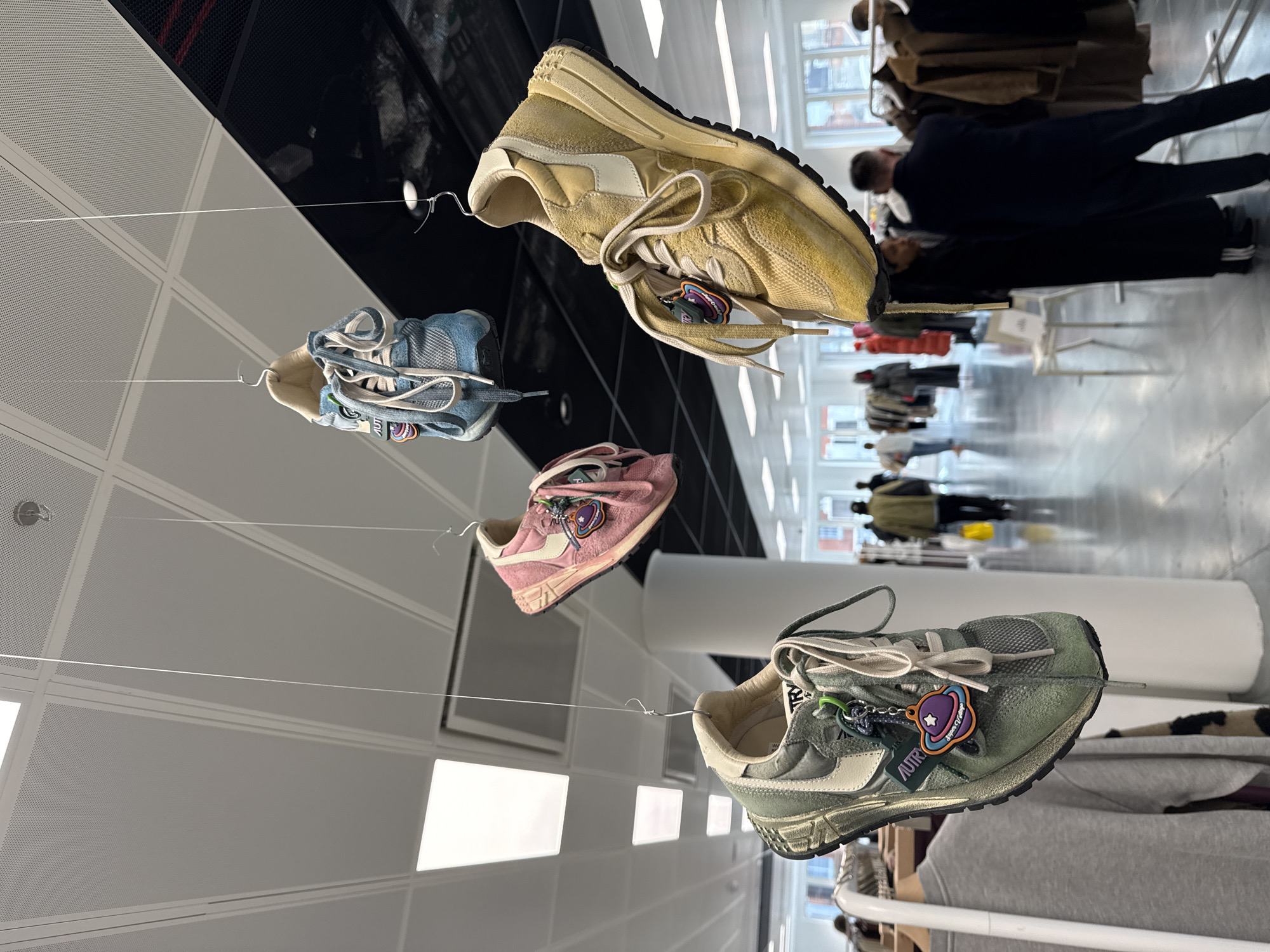 The image depicts an indoor exhibition in London, United Kingdom, featuring several pairs of sneakers suspended from the ceiling by thin wires. The sneakers are in varying colors and styles. The background features a modern interior with white walls, reflective floors, and large windows through which people and buildings can be seen. There are several people visible in the background, appearing to be visitors or staff. The lighting is bright and even, illuminating the sneakers and the surrounding environment.