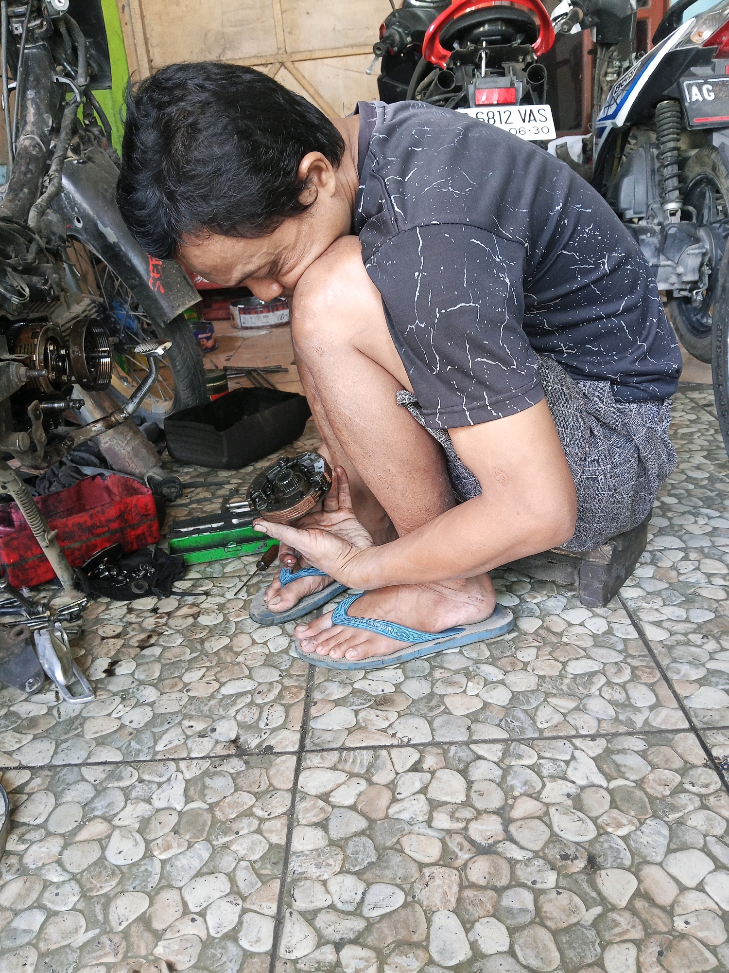 A man is kneeling on a stone-tiled floor next to a motorcycle, holding a disassembled motorcycle part in his hands. He is wearing a black t-shirt with a white splatter pattern and patterned shorts. Tools and other motorcycle parts are scattered around him. The scene appears to be an outdoor workshop or repair area. The motorcycles in the background indicate the setting could be in the city of Wates, Indonesia, a region known for its motorcycle culture and repair shops. The man is focused on his work, with his head bent low.