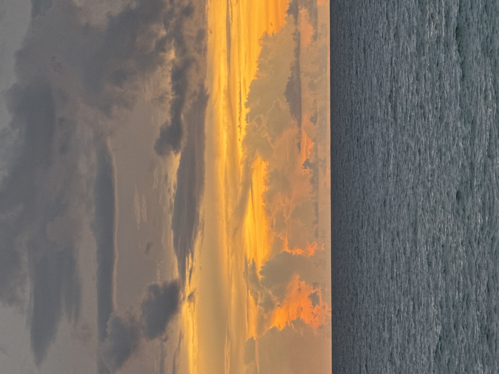An overcast sky is lit by the orange and yellow hues of a sunset over a dark, choppy ocean. The clouds are a mix of grays, with streaks of bright orange and yellow visible where the sunlight is breaking through. The ocean is a deep blue-gray with small waves and whitecaps. The horizon line is sharp and divides the sky from the sea. The scene is devoid of any human presence or man-made objects, suggesting a natural, isolated setting.