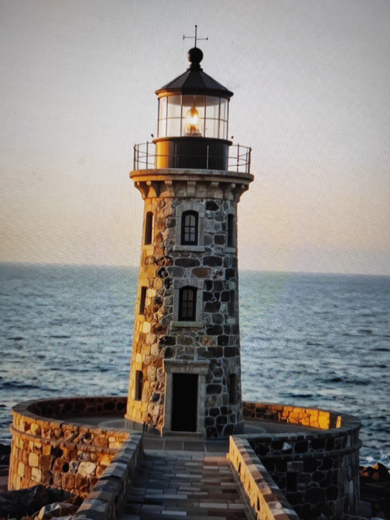 A stone lighthouse stands on a rocky shore, surrounded by water. The lighthouse is tall and cylindrical, with a conical roof and a bright light at the top. A walkway made of stone leads to the entrance of the lighthouse. The sky is a pale orange, and the water is a deep blue. The scene appears to be at sunset or sunrise.