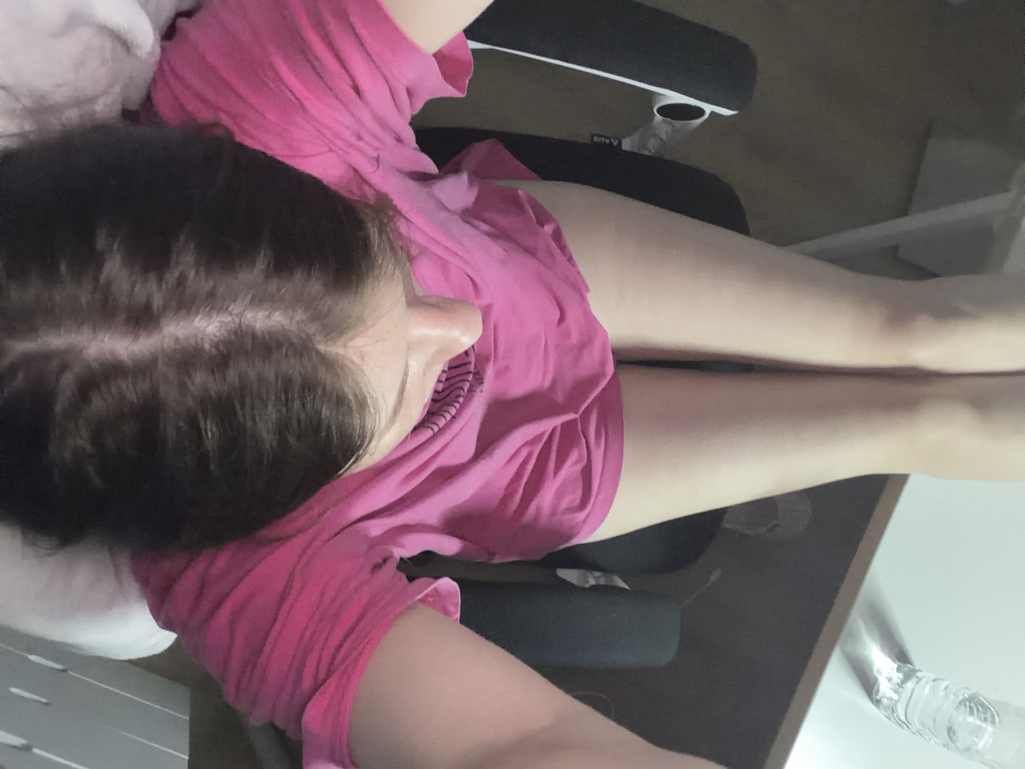 A young person is seated in a chair, viewed from directly overhead. The person has dark brown hair and is wearing a bright pink t-shirt. Their legs are visible, crossed at the thighs, and appear pale. The background shows a white pillow or headrest, and parts of a black chair. To the lower left, a desk or table is visible, with a clear plastic bottle of water on it. The overall setting appears to be indoors, possibly a home or office environment. There is no specific indicator of location within Dongducheon-si, South Korea.