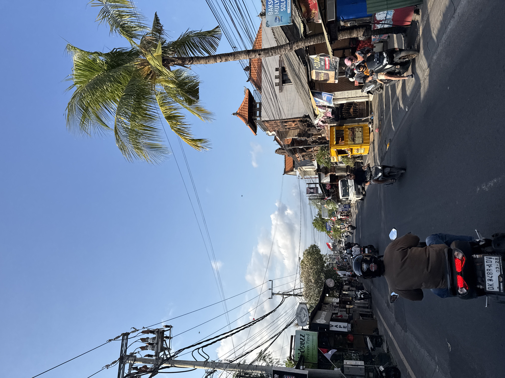 This image depicts a street scene in Denpasar, Indonesia, likely on Jalan Gajah Mada, known for its vibrant commercial activity. A person is riding a scooter in the foreground, seen from behind, wearing a brown shirt and blue jeans, with a black helmet. The scooter has a license plate that reads "DK 3488 ADV". The street is lined with shops and businesses, some with awnings and signs. One sign on the left reads "deuze Frozen Food". A tall palm tree with green fronds is visible on the right side of the frame, reaching towards a clear blue sky with scattered clouds. Several other scooters and motorbikes are on the road, some with passengers. The architecture features traditional Balinese elements, such as terracotta tiled roofs. Power lines are strung overhead, characteristic of urban infrastructure in the region. The overall atmosphere suggests a busy day in a tropical city.