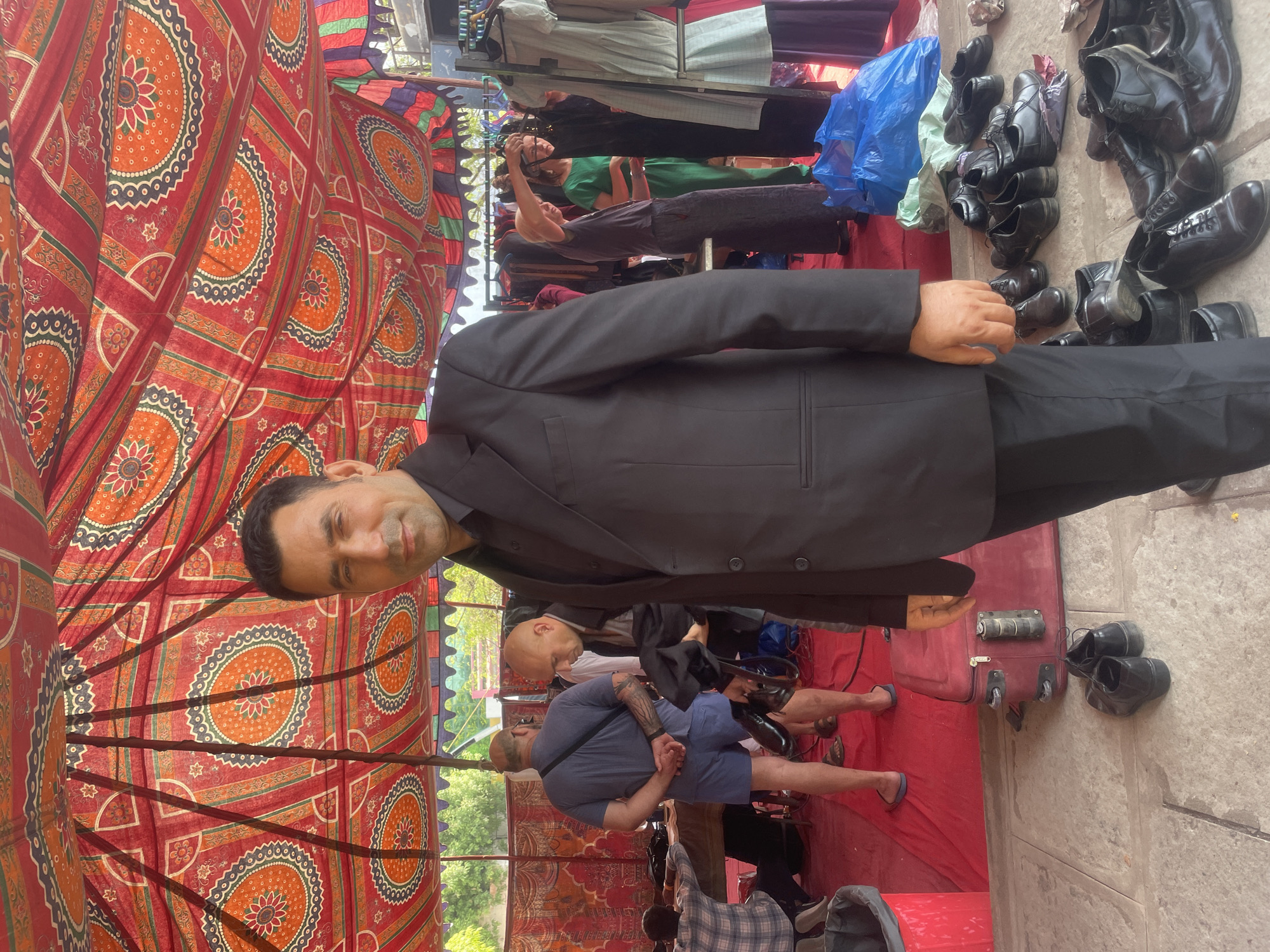The image captures a bustling market scene in Anajpur, India. A man in a black suit stands prominently, with stacks of black leather shoes arranged to his right. Behind him, a vendor tends to hanging clothes. Other people are visible in the background. The entire scene is covered by a colorful, patterned tent, offering shade.