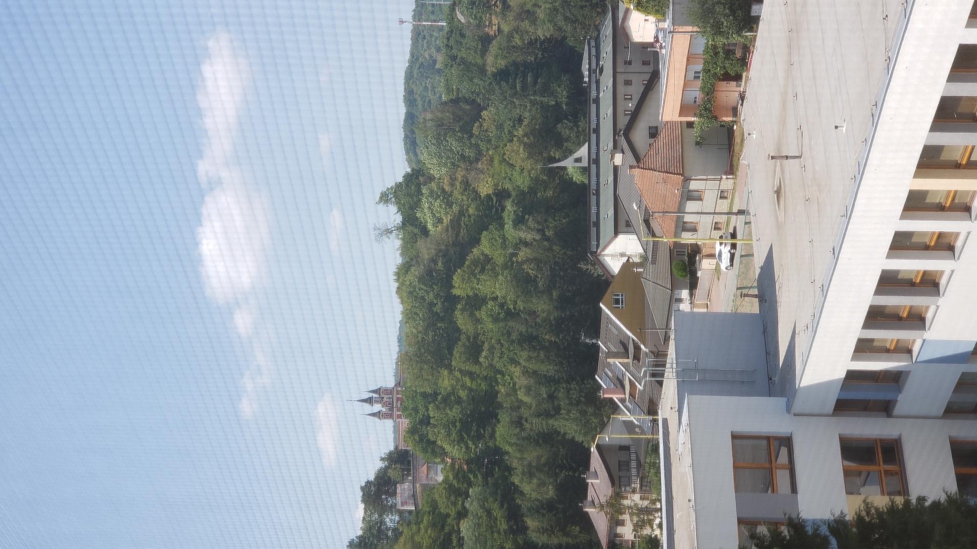 The image captures a view of a residential area and a wooded hillside in Prešov, Slovakia. In the foreground, a modern, multi-story building with a white facade and numerous windows is visible. Its flat roof is equipped with several ventilation pipes and anchoring points. Beyond this building, a collection of houses with various roof types, including red tiles and solar panels, are situated. A white car is parked on a paved street.
Dominating the background is a densely wooded hill, characteristic of the landscape surrounding Prešov. At the crest of the hill, a distinctive red and white building complex with multiple towers, likely a church or monastery, is prominently featured. The sky above is a clear, pale blue, with a few scattered white clouds. The overall scene depicts a daytime view of a town nestled against a natural, forested backdrop.
